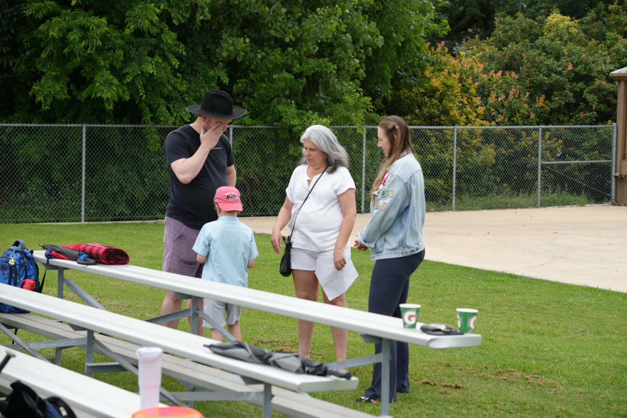 A group of four people, two women and two children, standing outdoors near a chain-link fence with trees in the background. One of the women has gray hair and is wearing a white shirt, the other has brown hair and is wearing a denim jacket. One child is wearing a pink hat, and the other has his head bent down. There are tables with drinks and backpacks on them in the foreground.