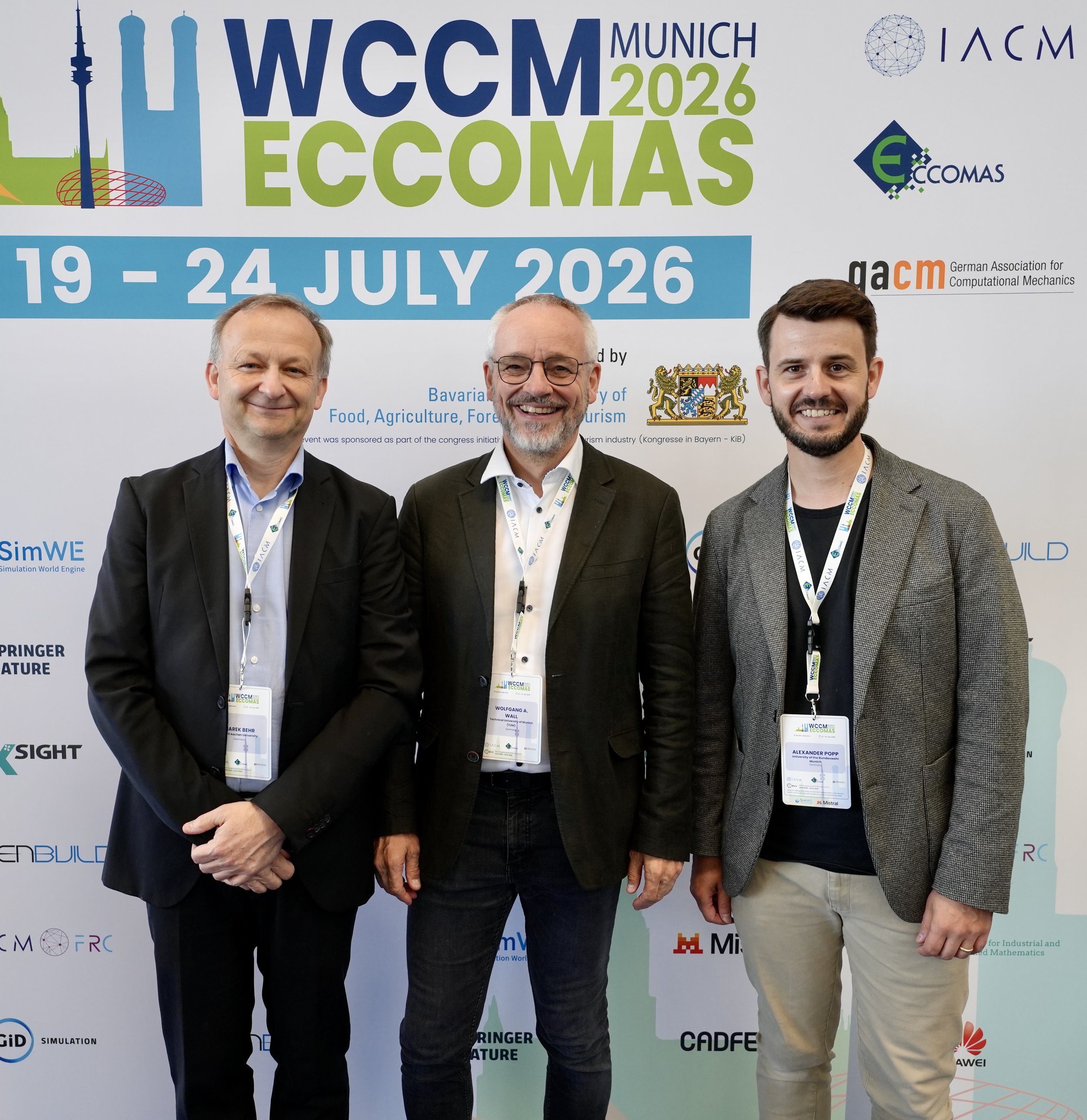 Three men standing in front of a conference banner with the text "WCCM Munich 2026 ECOMAS" and the dates "19-24 July 2026." The men are dressed in business attire, wearing conference lanyards and smiling at the camera. The background includes logos and text related to the conference and sponsors.