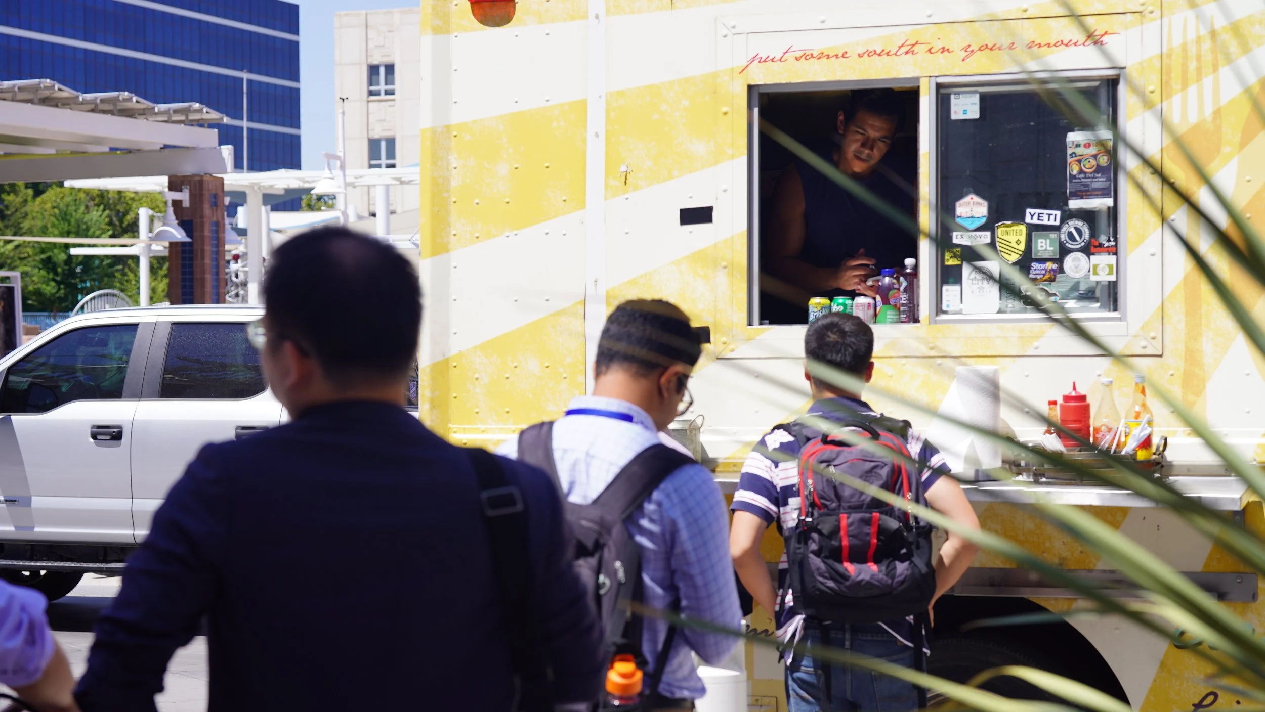 People standing in line at a yellow food truck, with one person inside preparing food, and a blue modern building in the background.