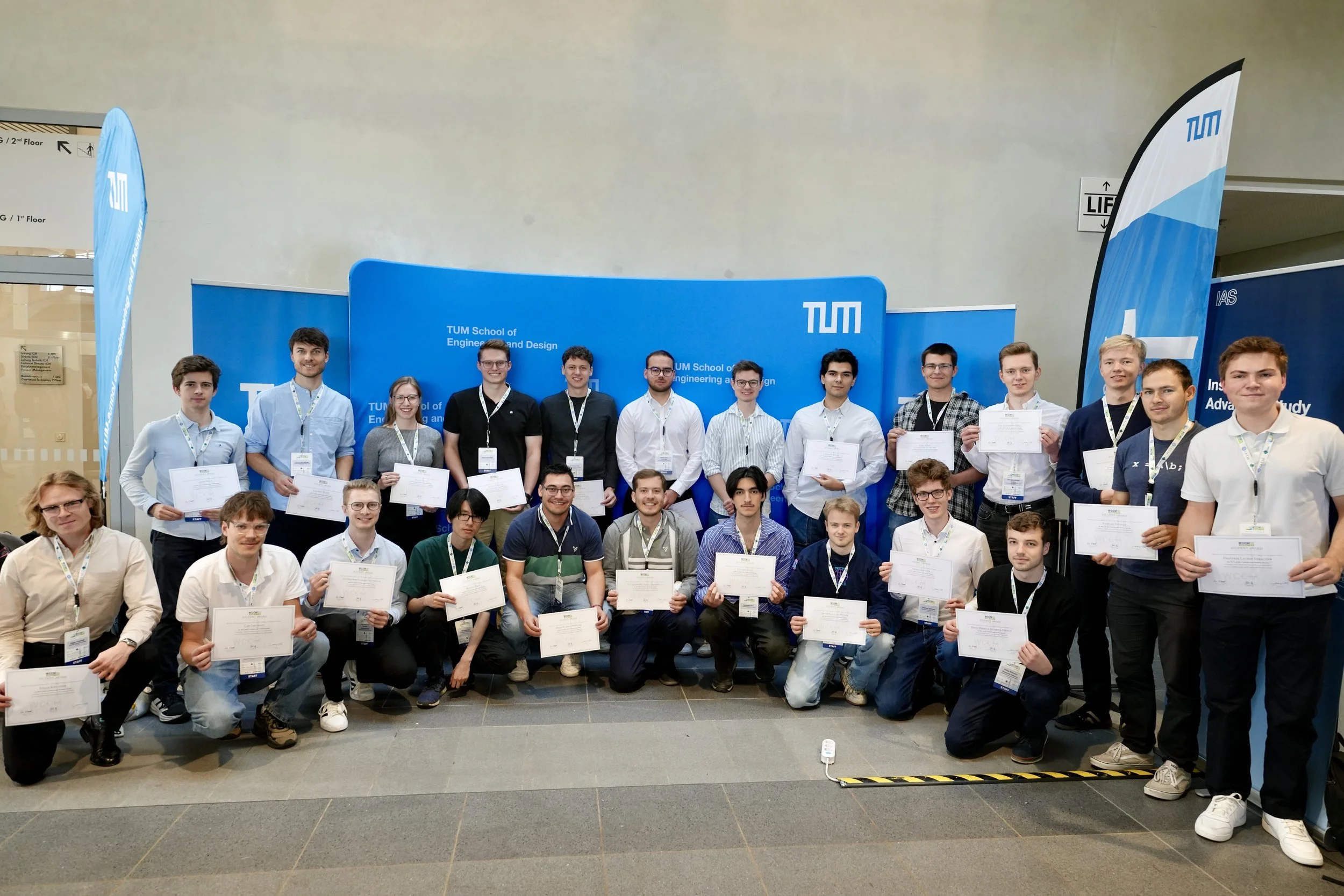 A group of students and young adults posing with certificates at a university event, with blue TUM (Technical University of Munich) banners in the background.