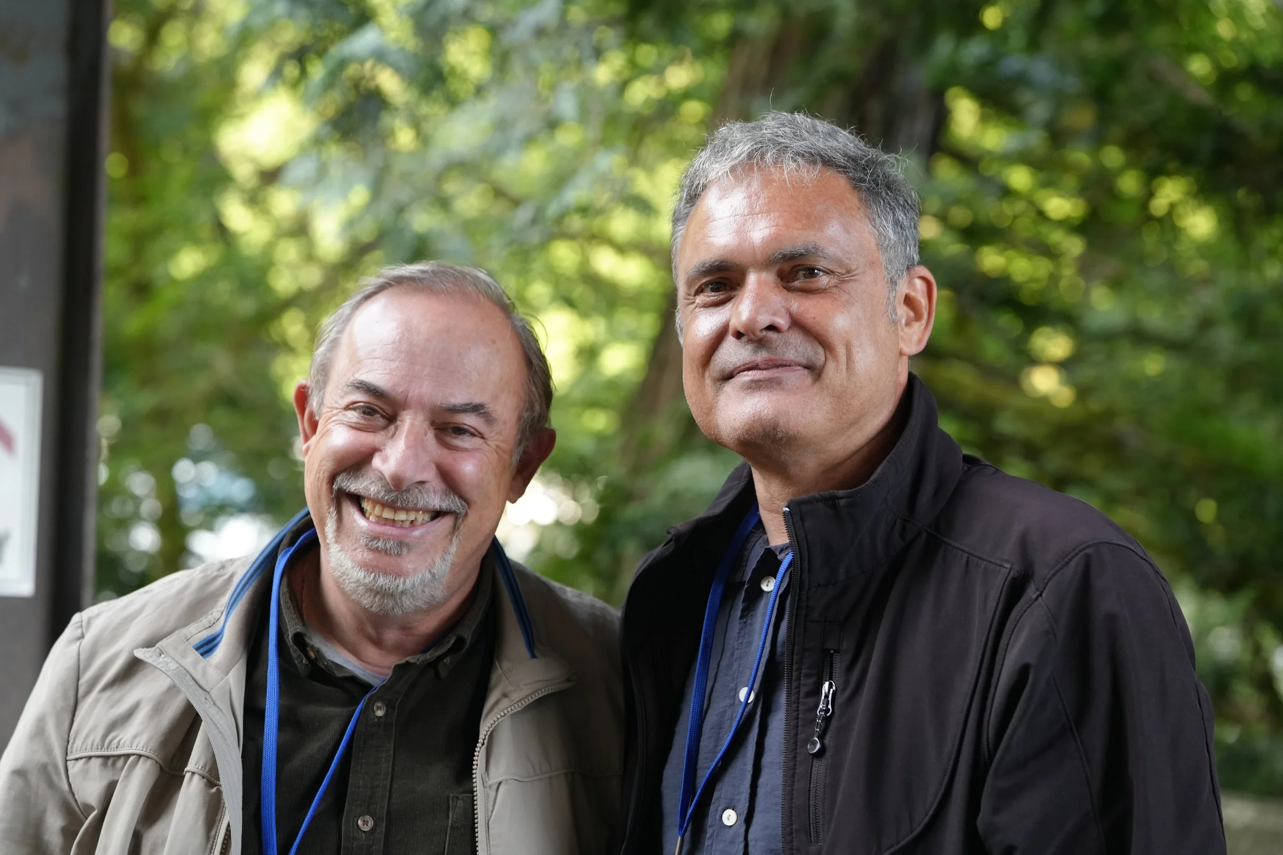 Two smiling middle-aged men standing outdoors in a green, leafy park, close together, in casual clothing.