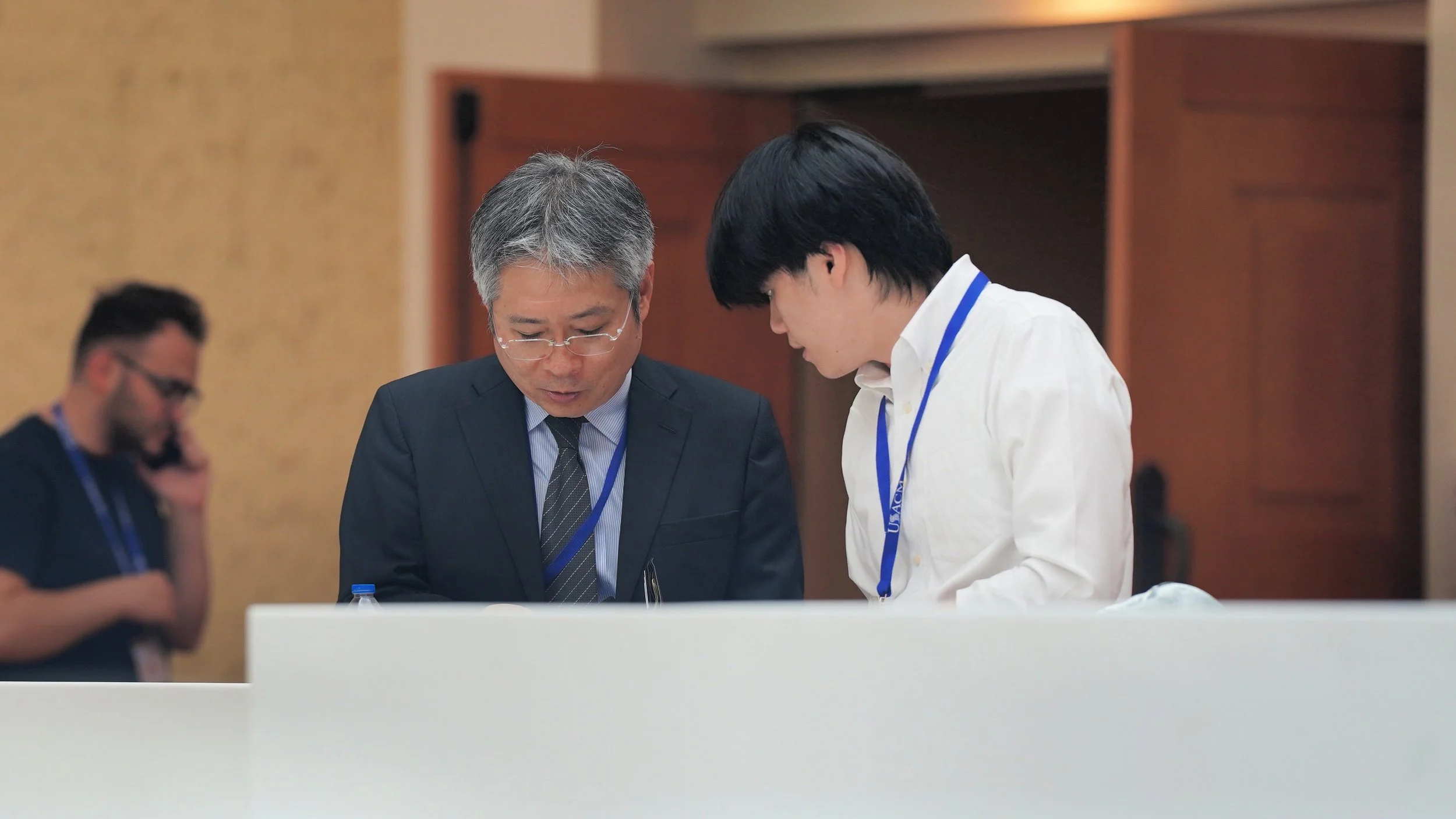 Two men engaged in conversation at a conference, one in a suit and the other in a white shirt, with conference badges around their necks.