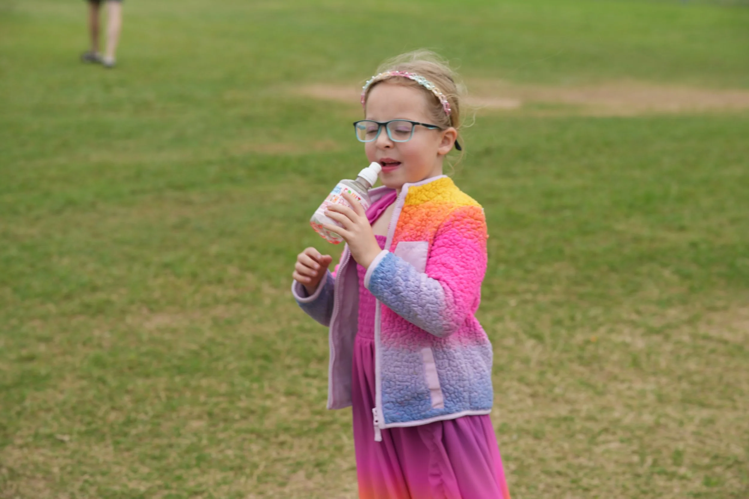 A young girl with glasses and a colorful fleece jacket drinking from a bottle on a grassy field.
