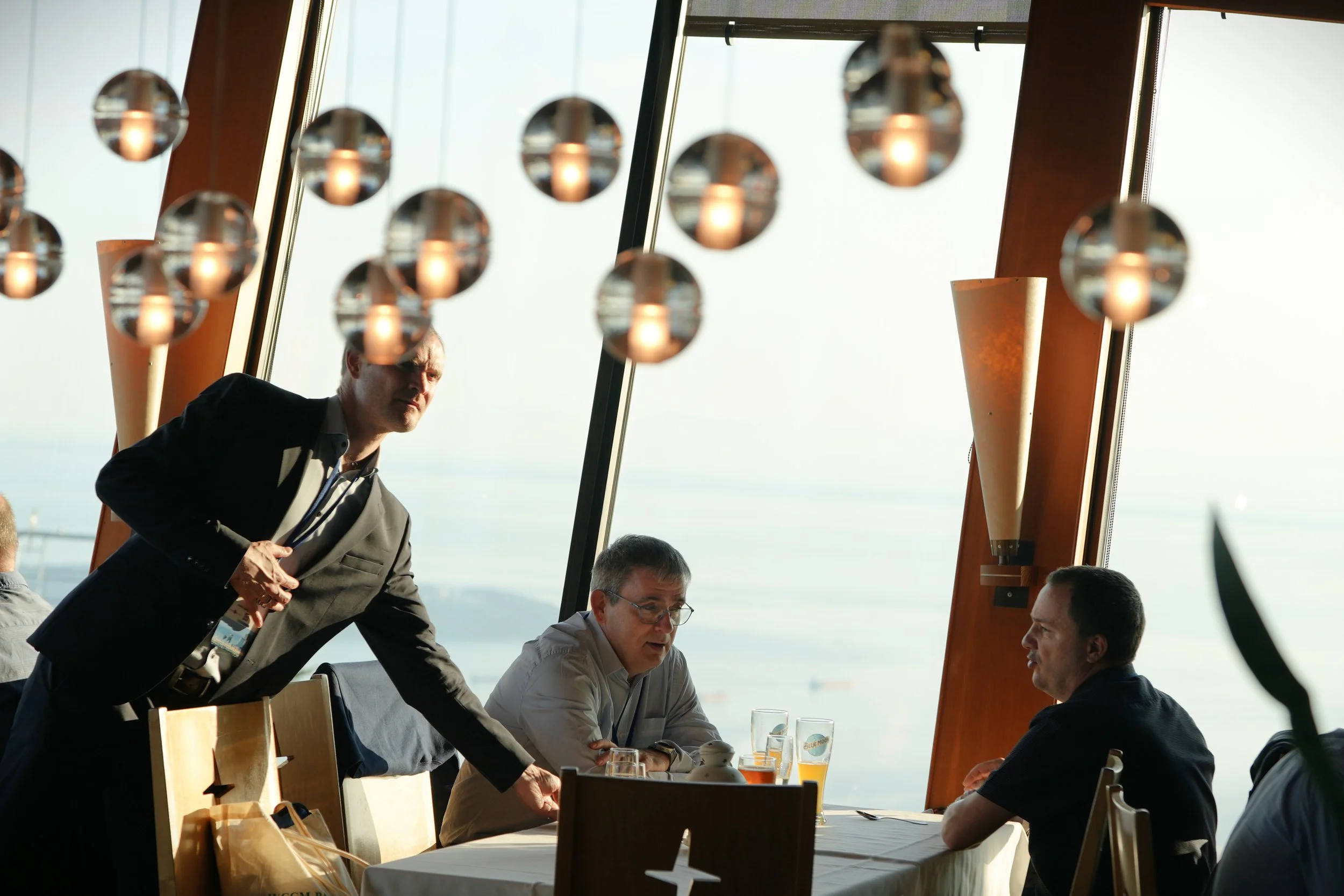 A man in a dark suit has a conversation with another man seated at a table, with a third person also present. The setting appears to be a restaurant or conference room with large windows showing an outside view and unique hanging lights.