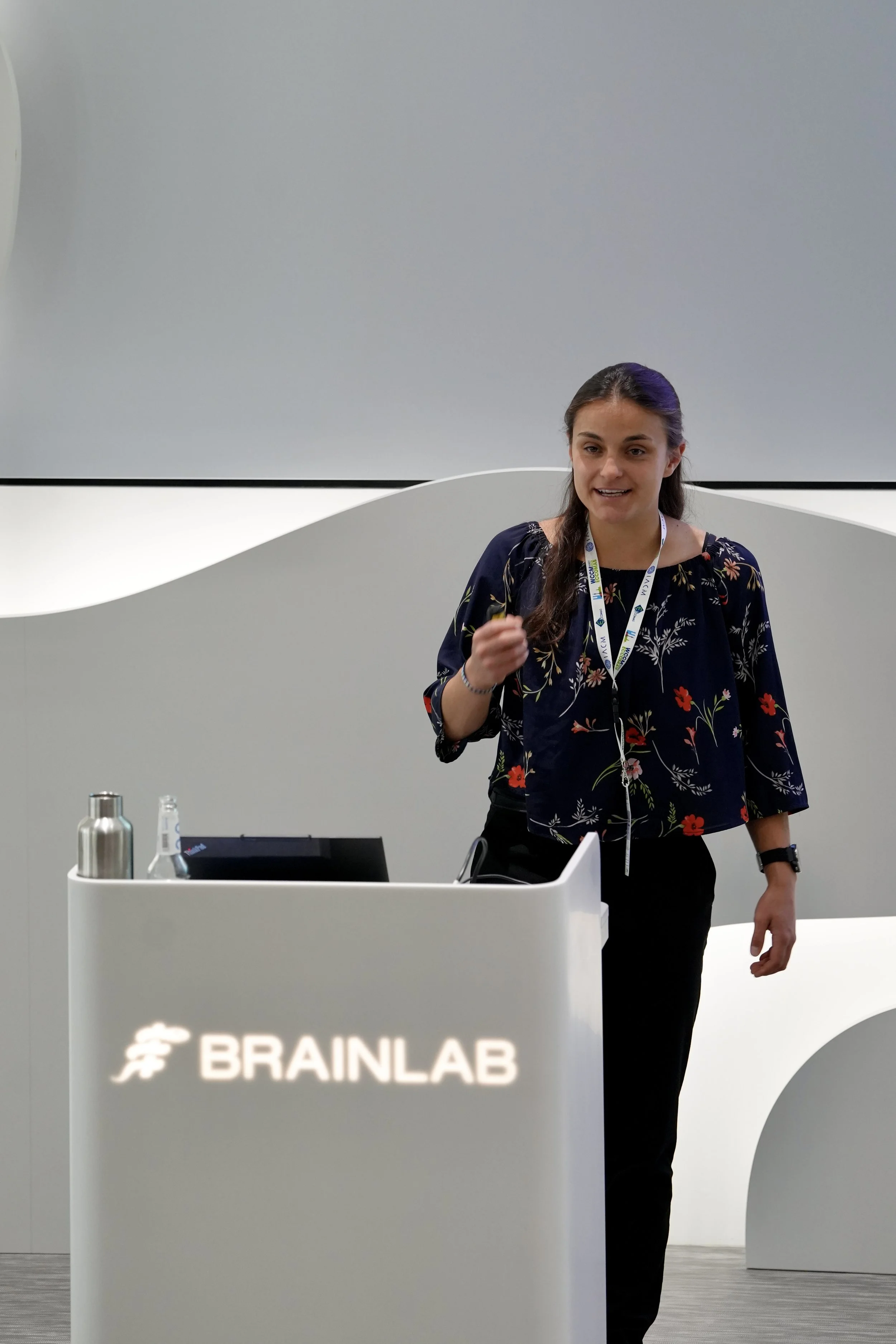 A woman standing behind a white podium with the '#BRAINLAB' logo, wearing a navy blue floral blouse and a lanyard, speaking or presenting at an event.