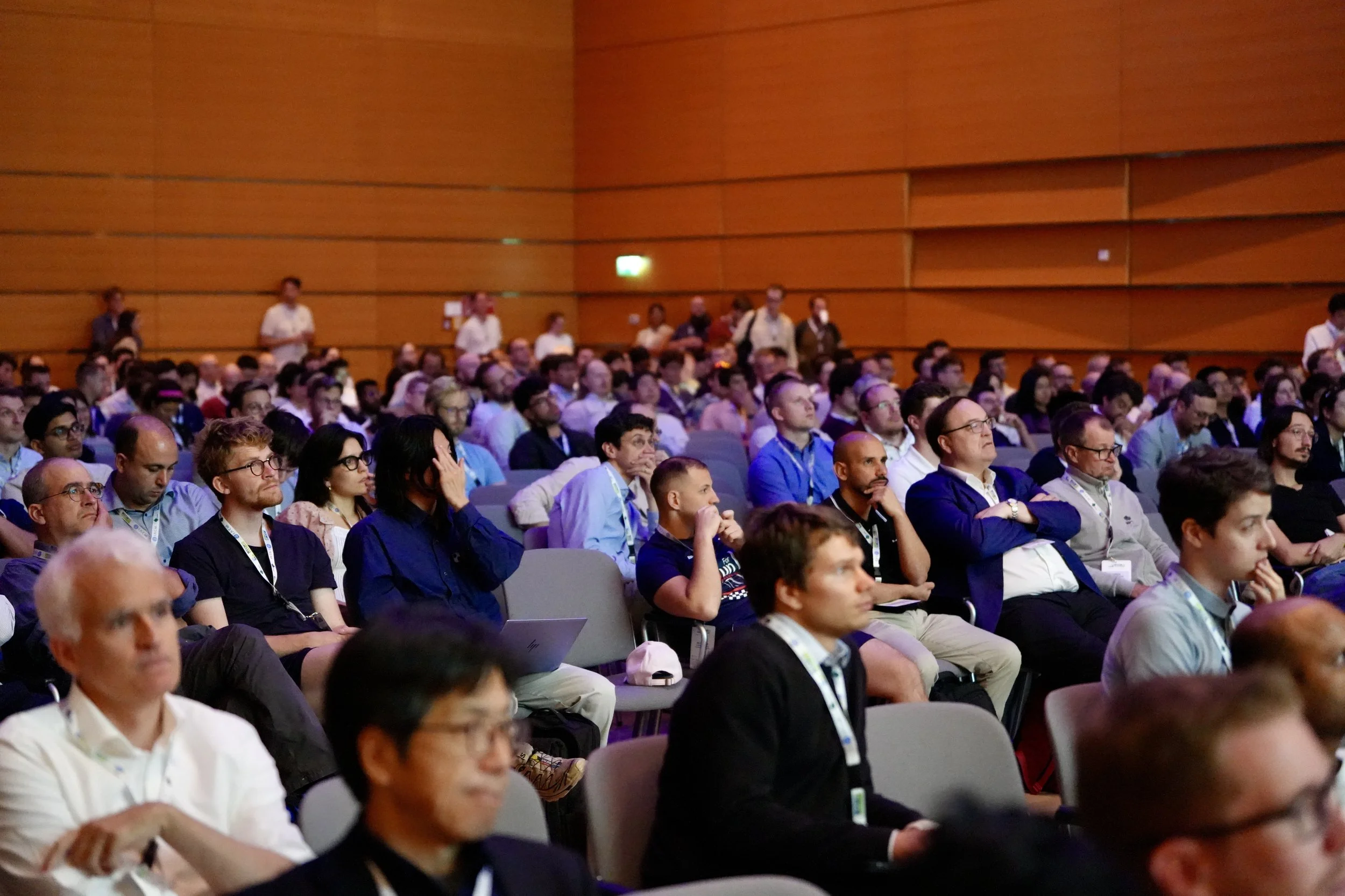 An audience of diverse people attending a conference or seminar in a large auditorium with wooden panel walls, many are focused on a speaker or presentation not visible in the image.