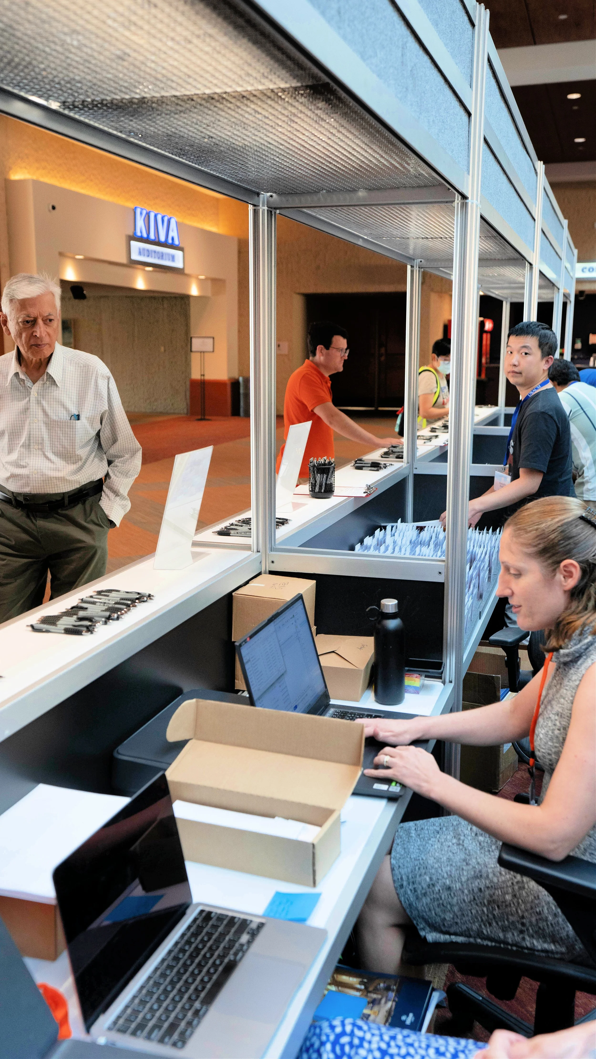 A registration or information booth at a conference or event with people attending, some seated at tables with laptops and others standing or walking nearby, in a large indoor venue with a sign for the Kiva Auditorium in the background.