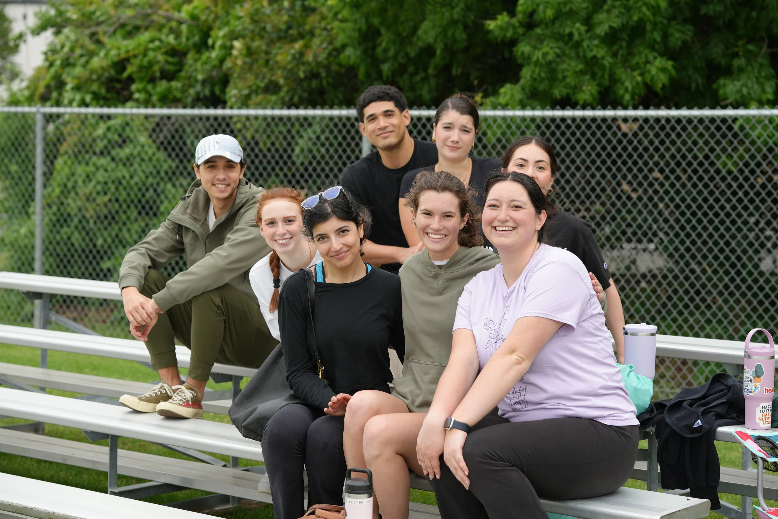 Group of nine diverse young people sitting on bleachers outdoors near a chain-link fence with greenery in the background.