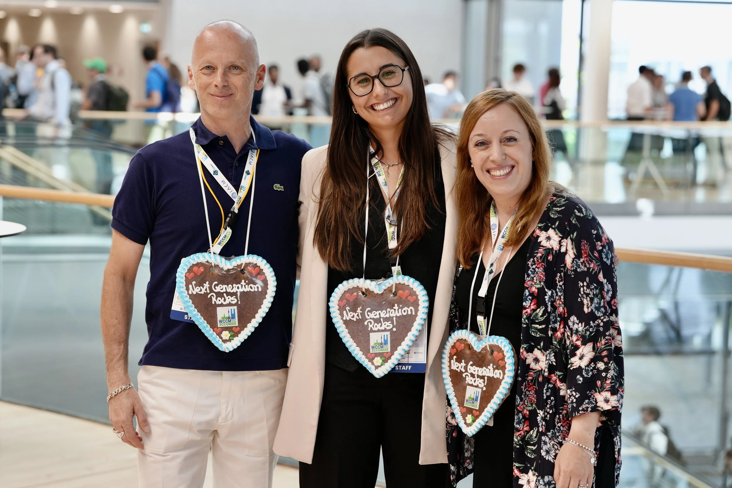Three smiling women wearing gingerbread heart necklaces with the phrase "Next Generation Rocks!" printed on them. They are at a conference or event with a busy background of people and a large glass window.