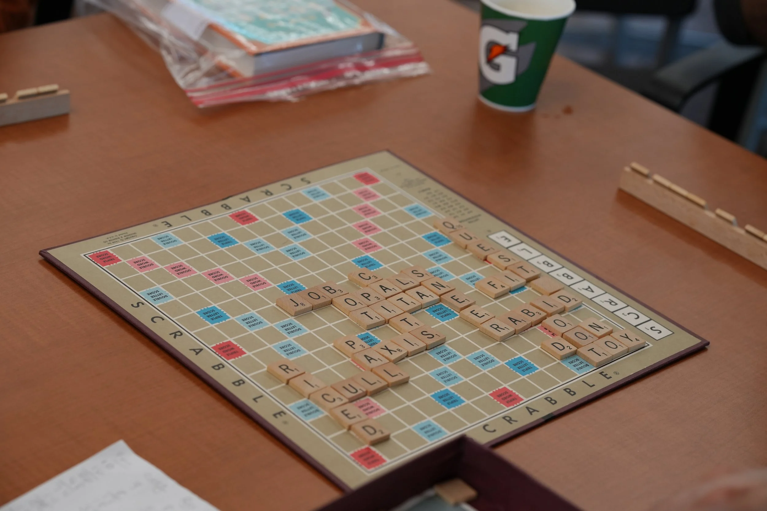 A Scrabble game in progress on a wooden table, with game tiles spelling words like 'JOULE' and 'PAL' and a Gatorade cup nearby.