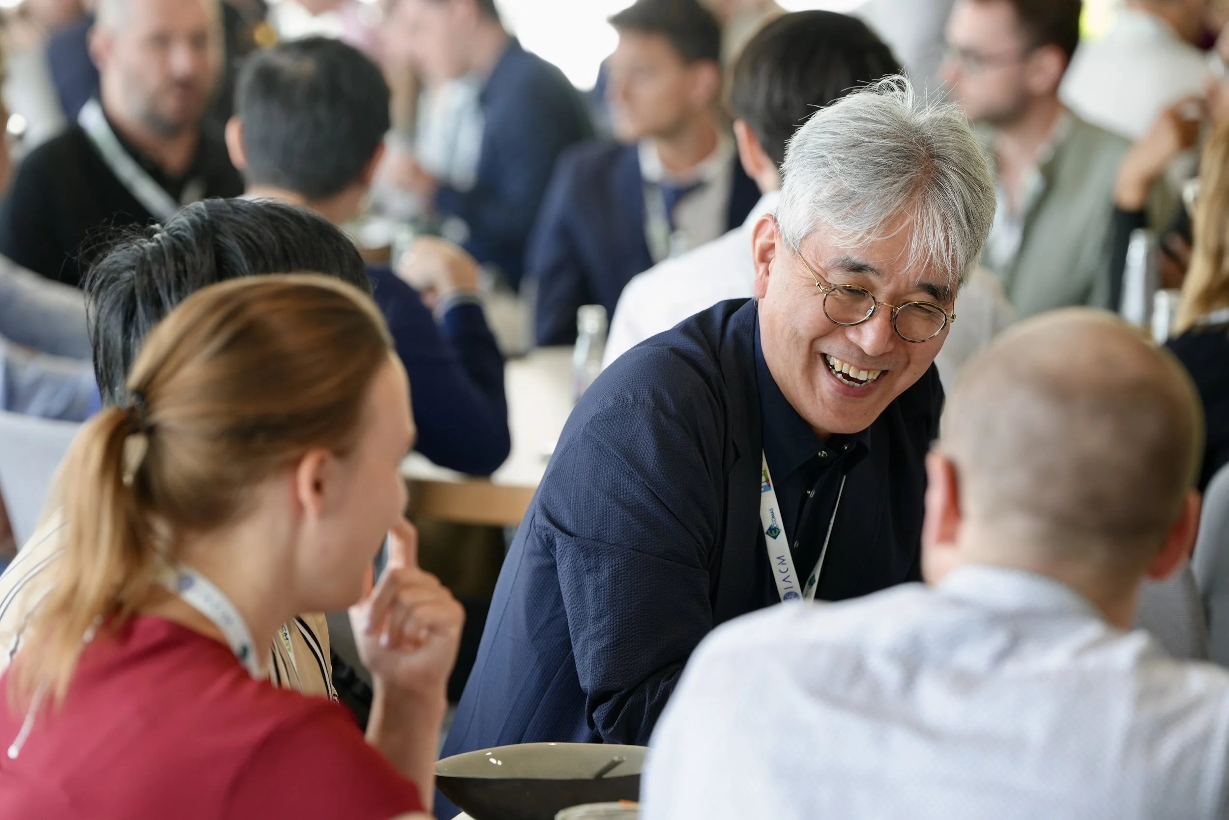A group of people sitting around a table at a social or professional event, engaged in conversation, with one older man smiling and wearing glasses in the center.