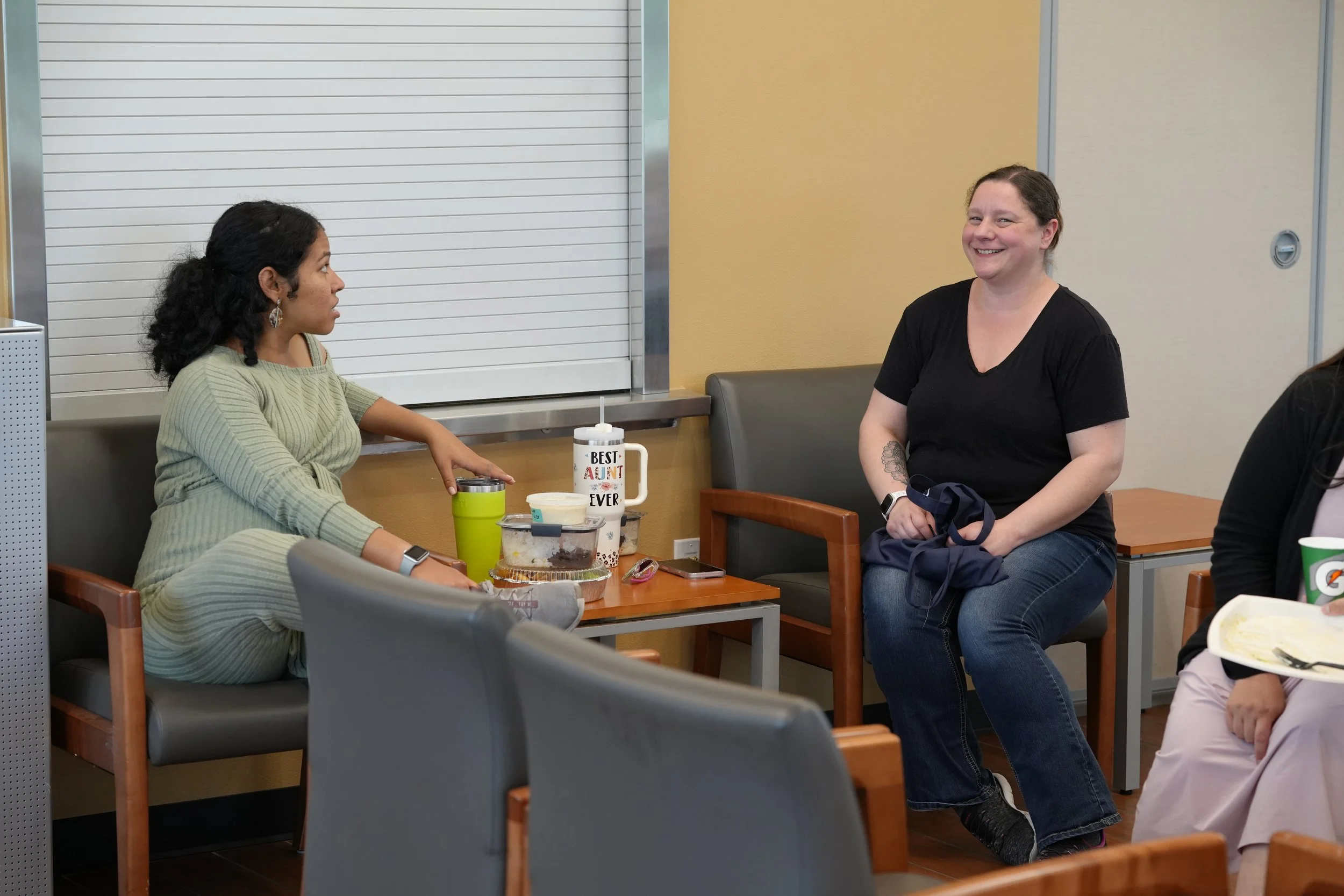 Two women sitting and conversing at a table with food and drinks, in a casual indoor setting with a closed metal shutter and beige wall behind them.
