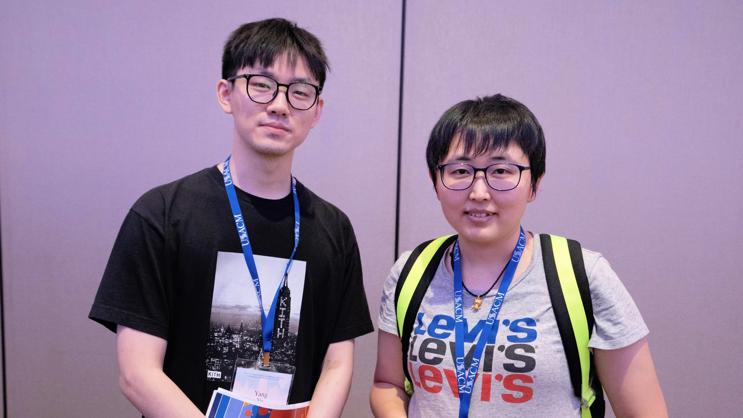 Two young men with glasses at a conference, wearing lanyards and casual t-shirts, standing against a purple wall.