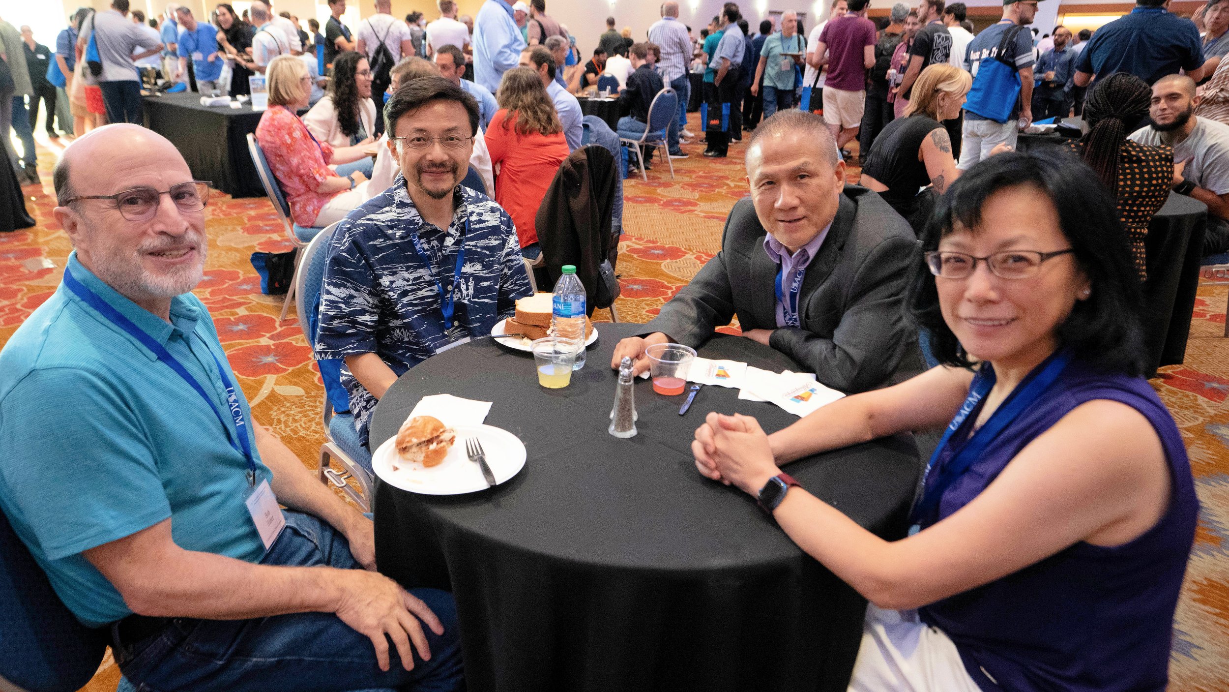 A diverse group of four people sitting around a table at a conference or event, with a crowded room of attendees in the background. They are smiling and appear to be enjoying the event. The table has plates with food, a bottle of water, and small cups with drinks.