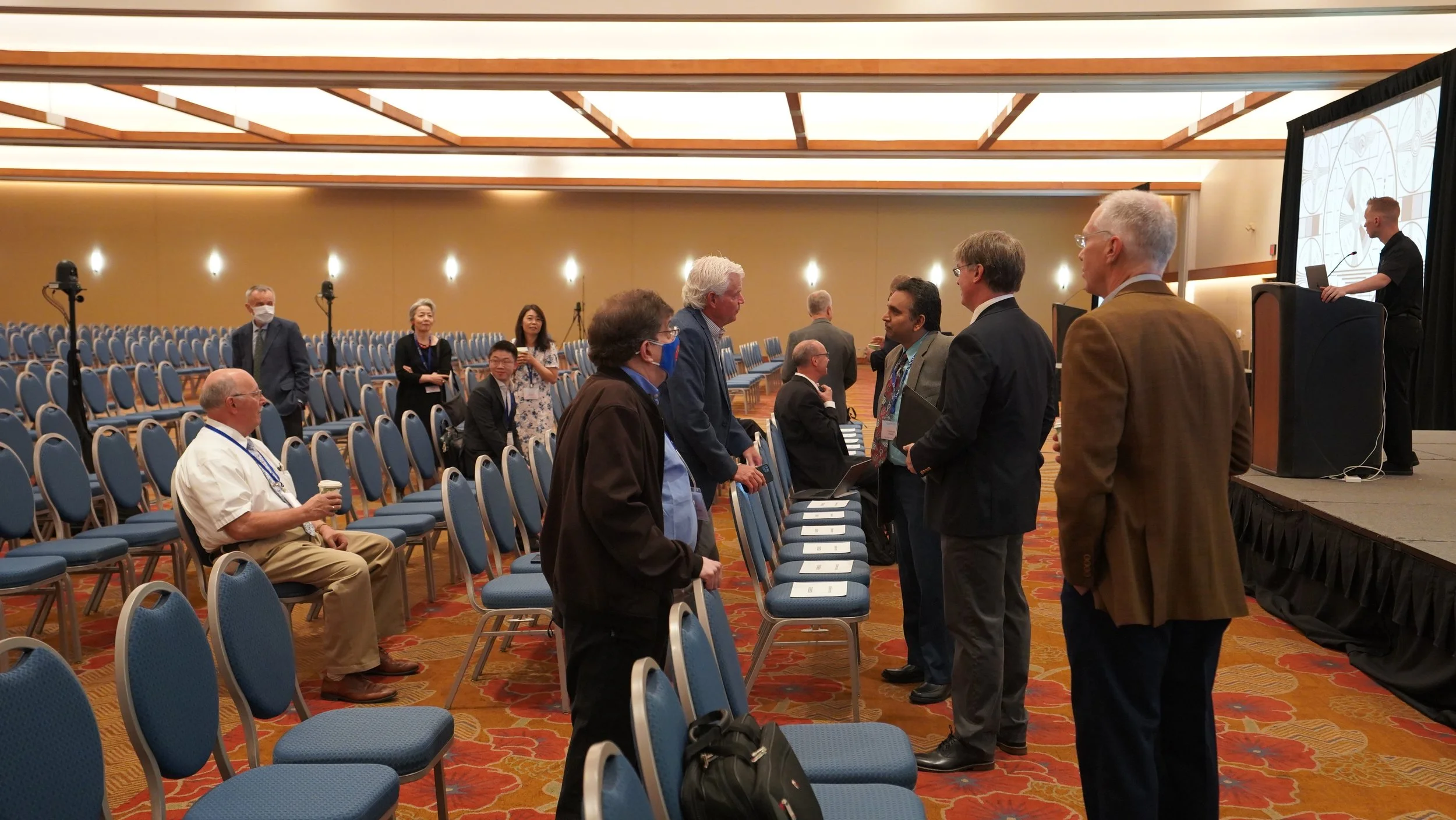 People attending a conference in a large, well-lit room with blue chairs and a stage. Some are seated, others are standing and talking, and a speaker is on the stage at a podium.