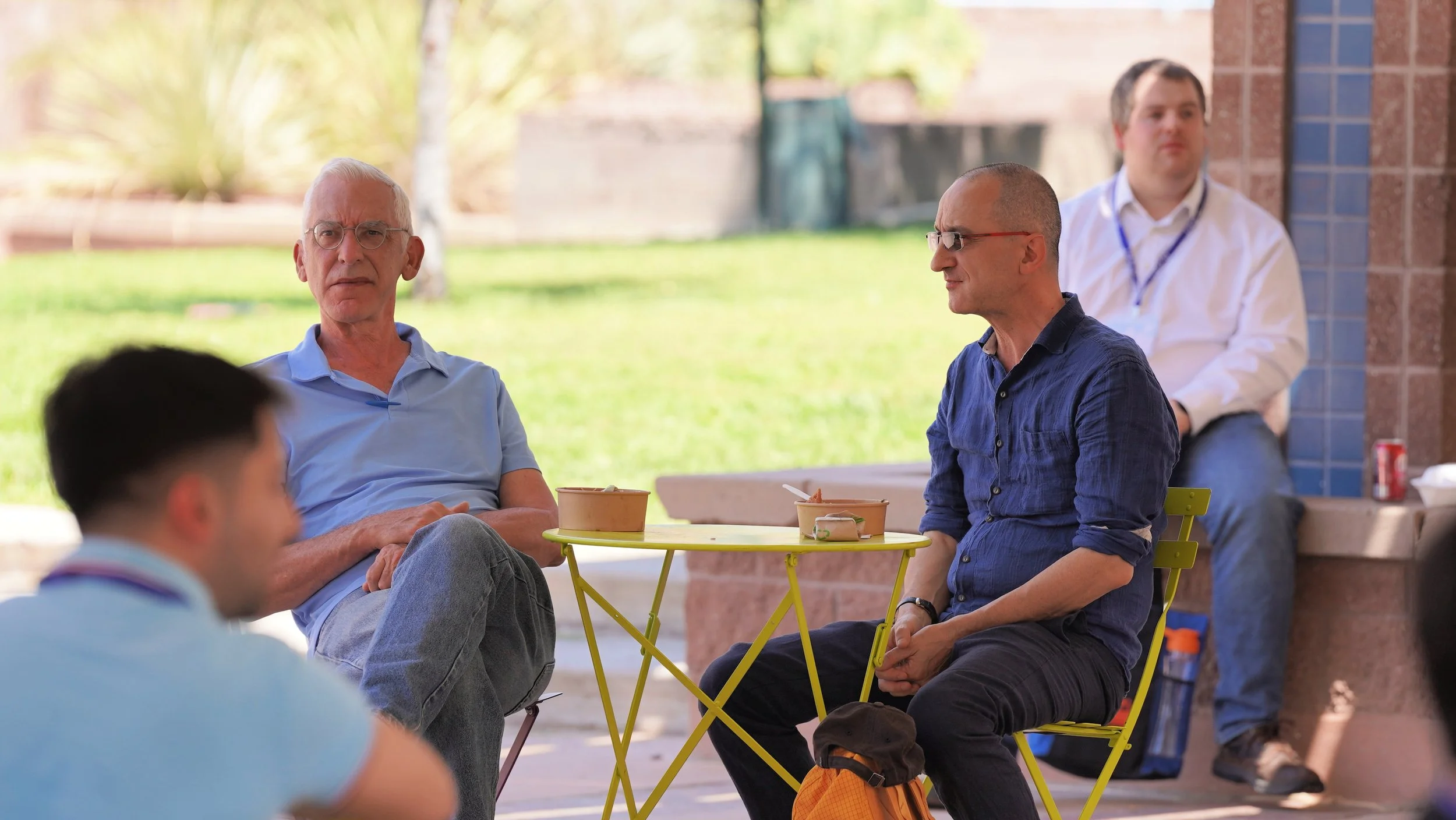 A group of men sitting outdoors at a yellow table, having a conversation. The background features trees and a brick wall.