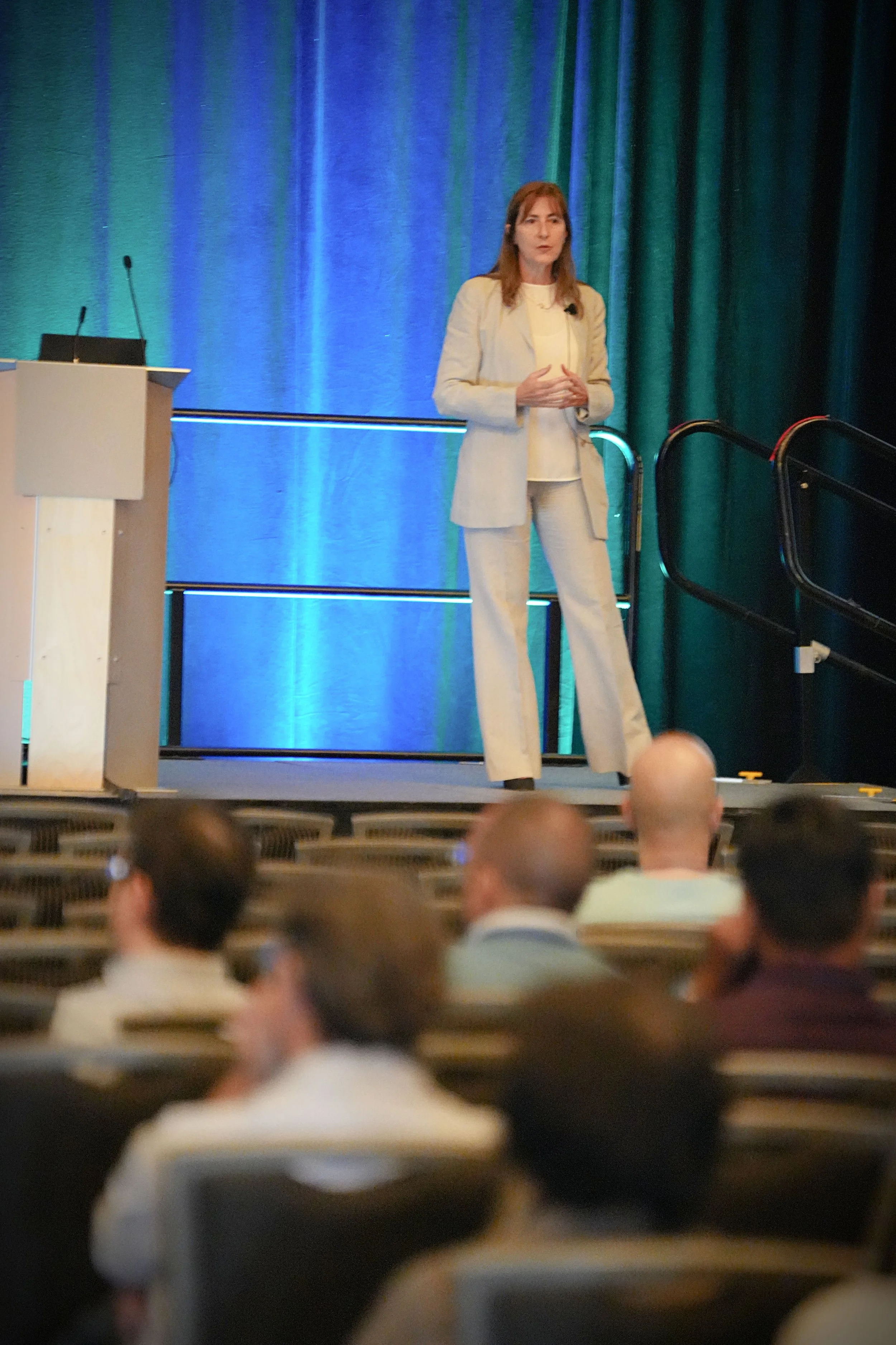 Woman giving a presentation on stage in front of a green and blue curtain, speaking to an audience in a conference room.