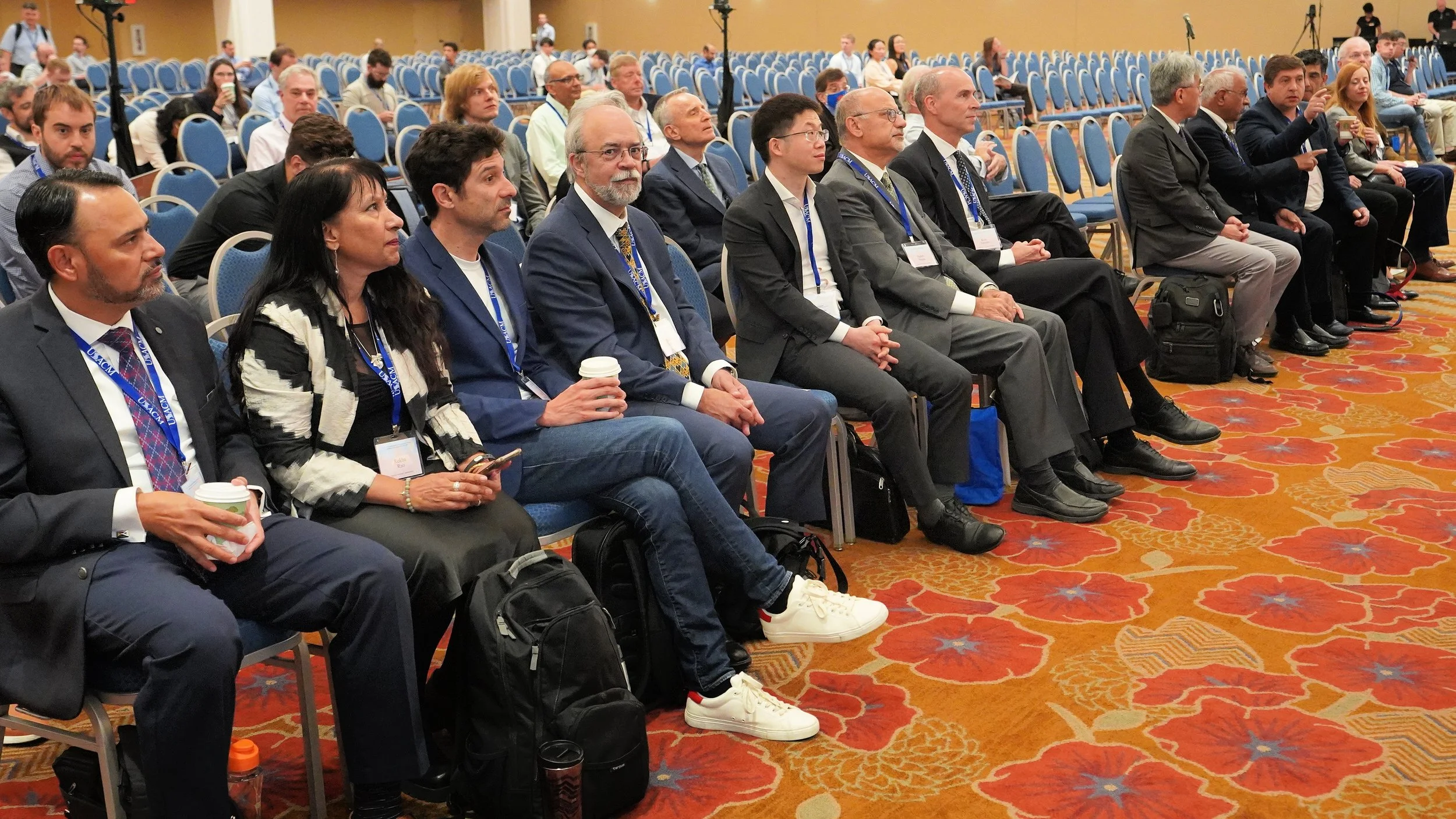 Attendees seated at a conference or seminar, some holding coffee cups, in a large room with blue chairs and a red floral-patterned carpet.