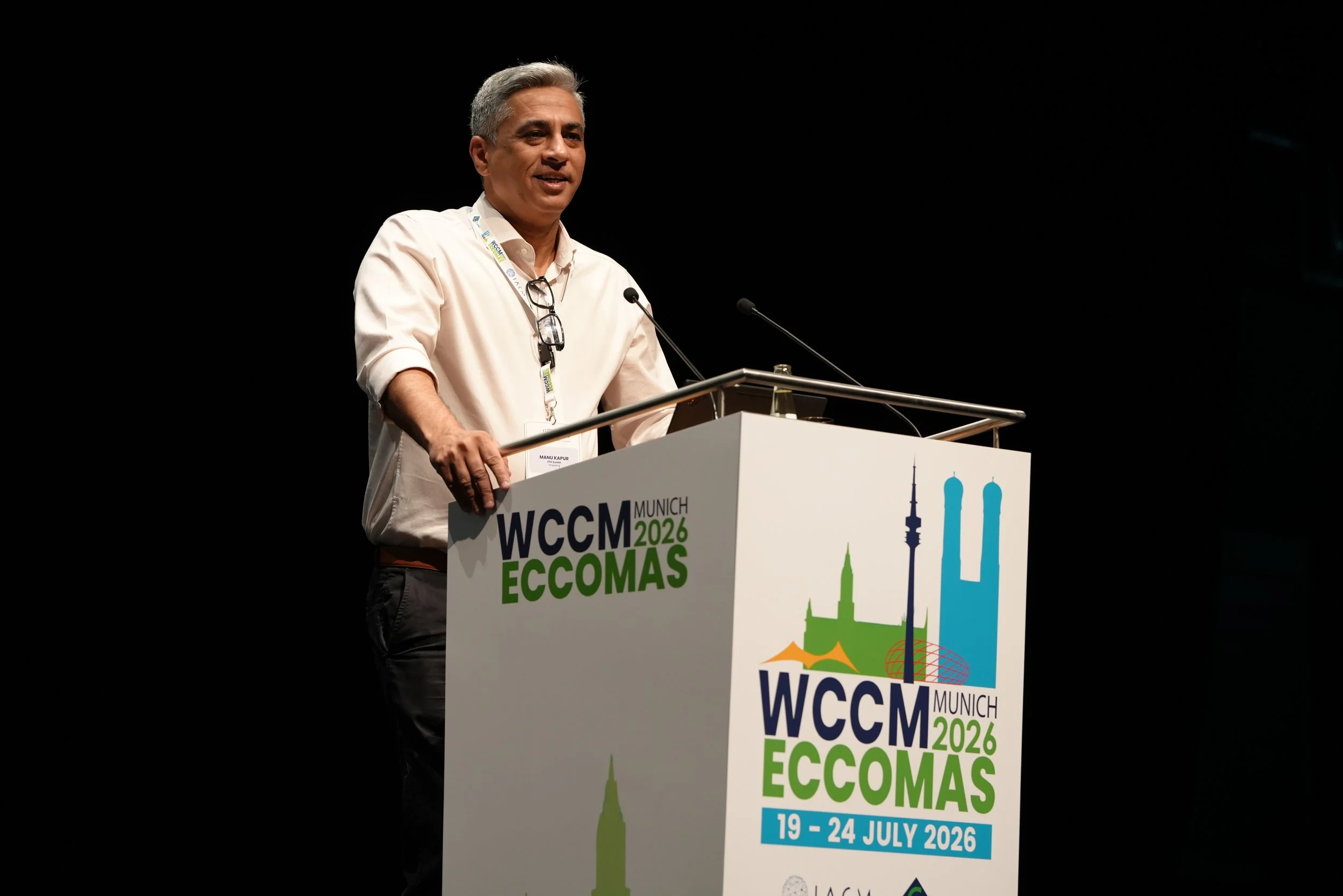 A man standing at a podium speaking during an event titled 'WCCM Munich 2026 ECCMAS' held from July 19-24, 2026. The podium has colorful graphics of famous structures and the event details.