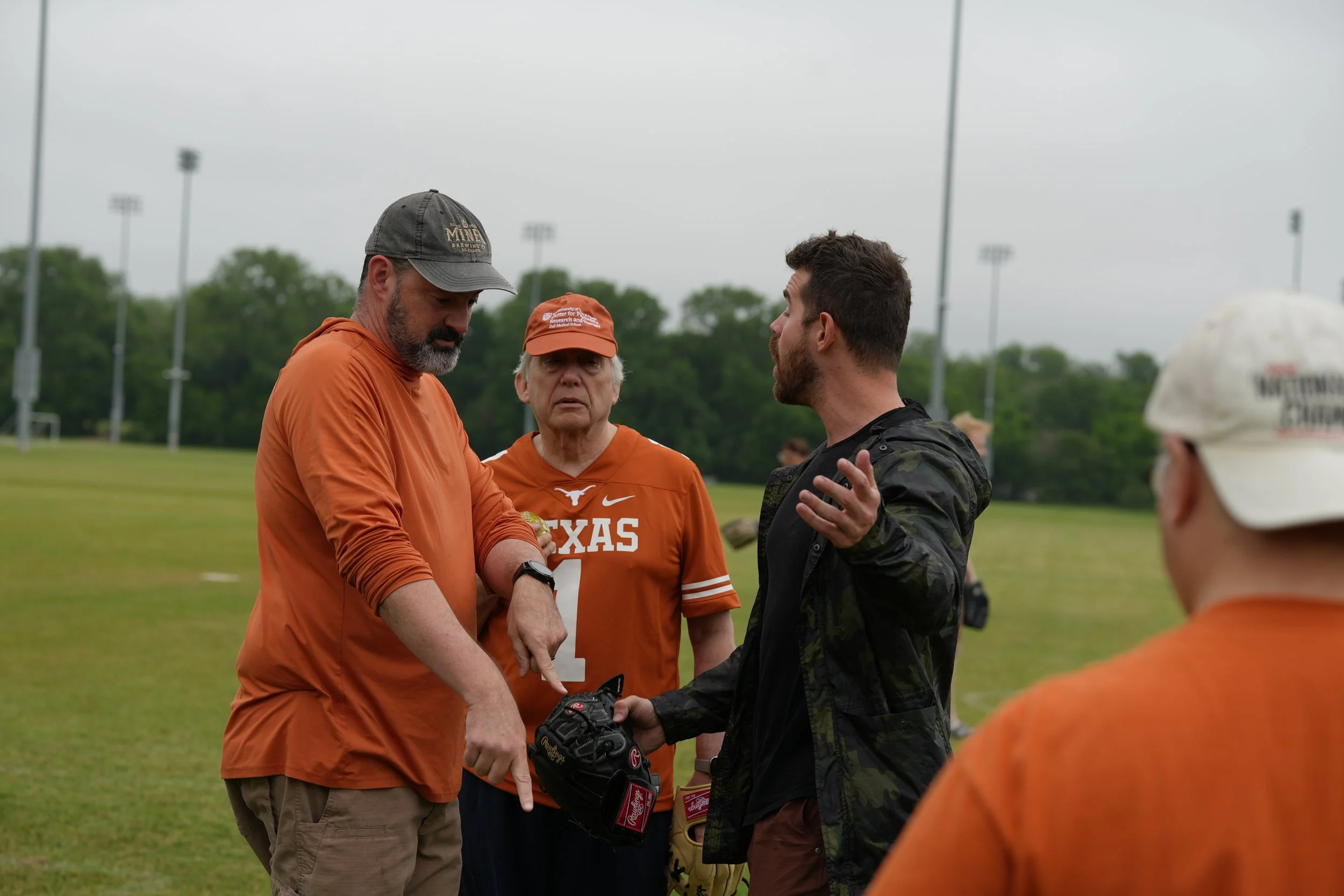 A group of people, including men in orange athletic jerseys and a man in black, are gathered on a sports field, engaging in conversation and handshakes under an overcast sky.