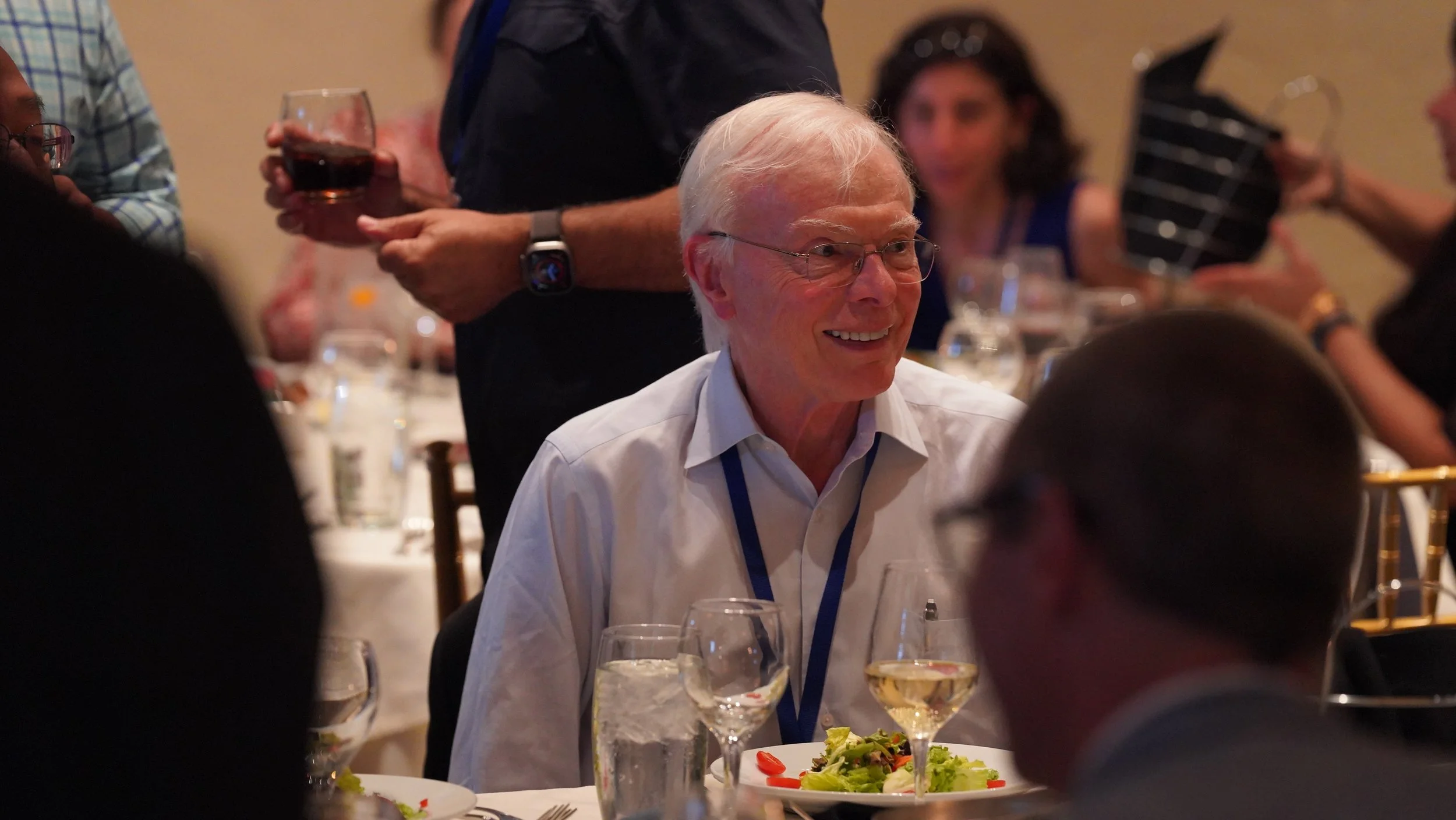 An elderly man with white hair and glasses smiling while seated at a dinner table during a social event, with a plate of salad and glasses of white wine in front of him, other people in the background, some holding wine glasses and a person with a tablet.
