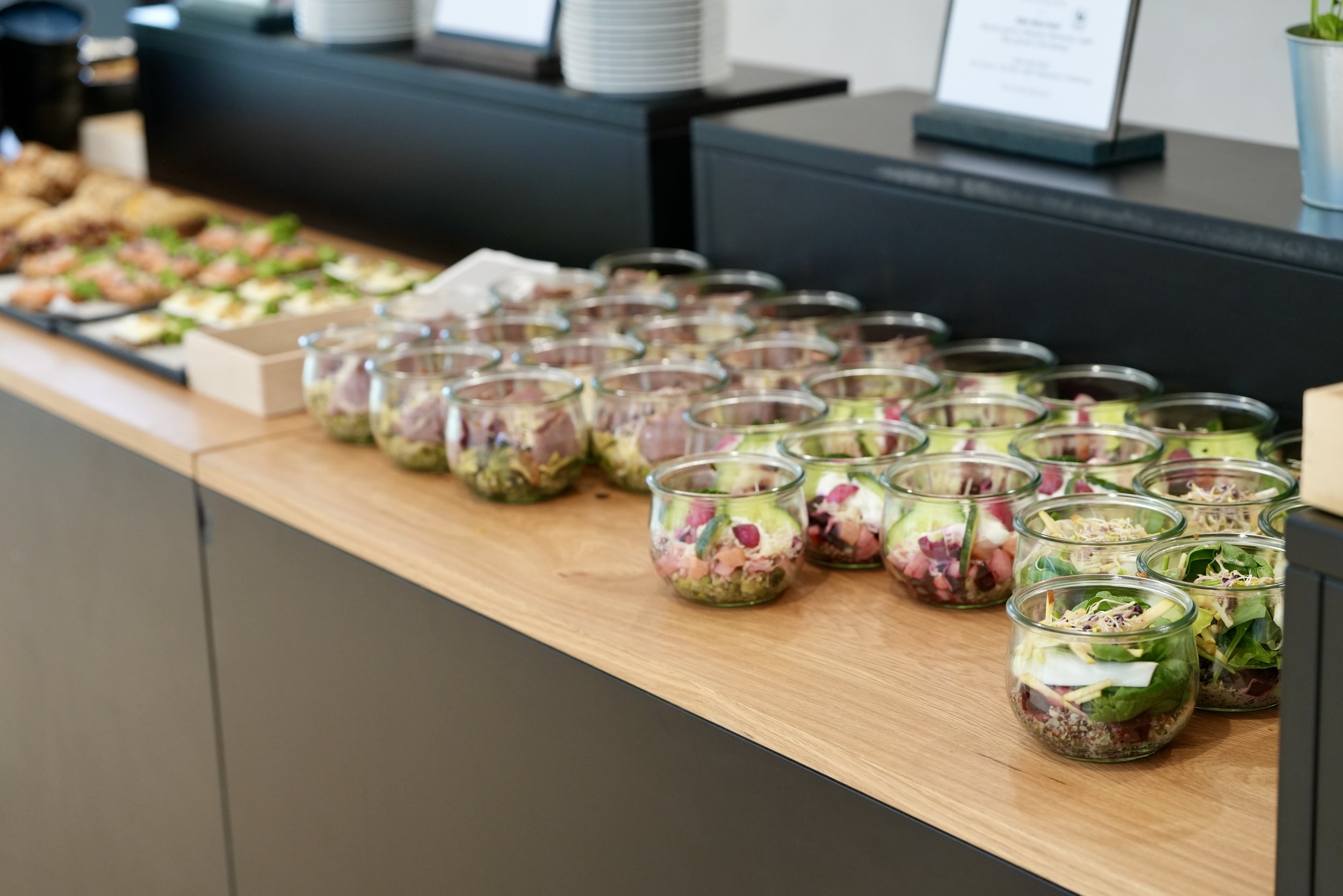A display of assorted salads and appetizers in small glass bowls on a wooden table in a restaurant or catering setting. These are conference snacks.