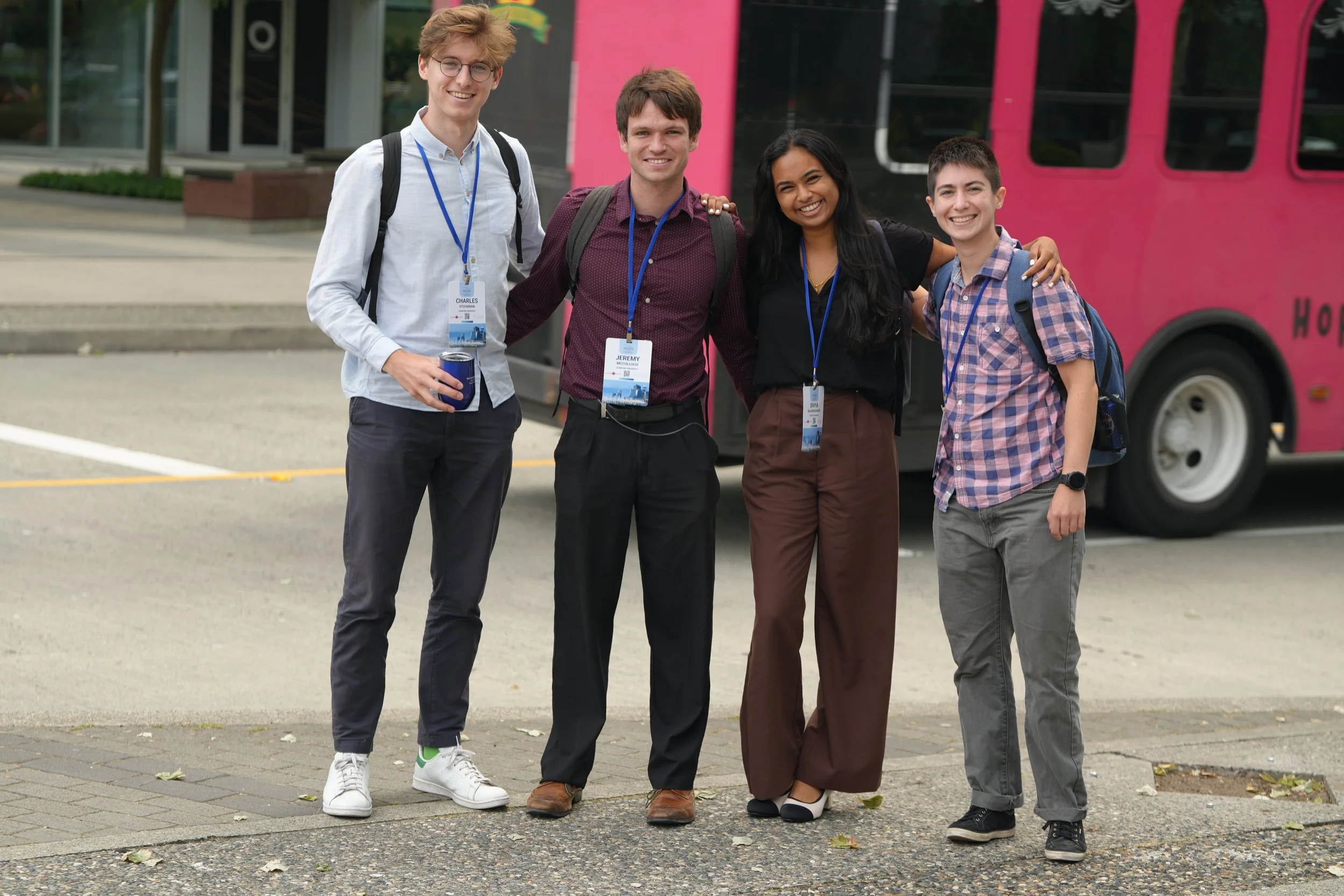 Four young adults, three men and one woman, smiling and posing together outdoors in front of a pink bus. They are wearing casual clothes, carrying backpacks and wearing conference or event badges.