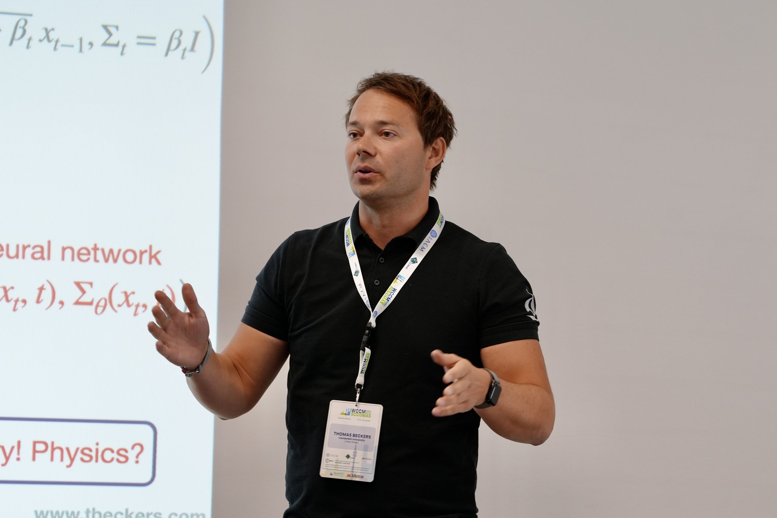 A man giving a presentation in front of a slide with mathematical formulas. The man is wearing a black polo shirt, a conference badge, and a watch, and appears to be explaining something.