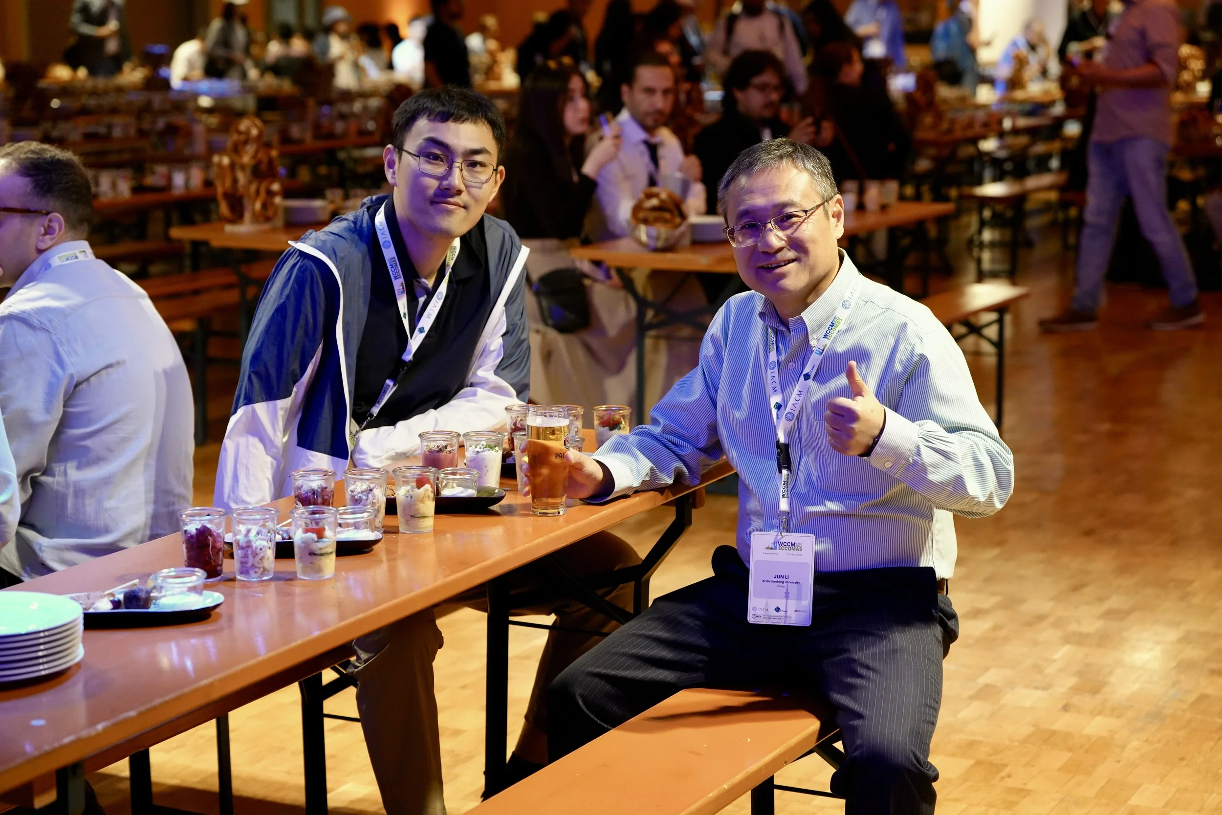 Two men sitting at a table during a food event, smiling at the camera, with others in the background at a crowded indoor venue with wooden tables and chairs.