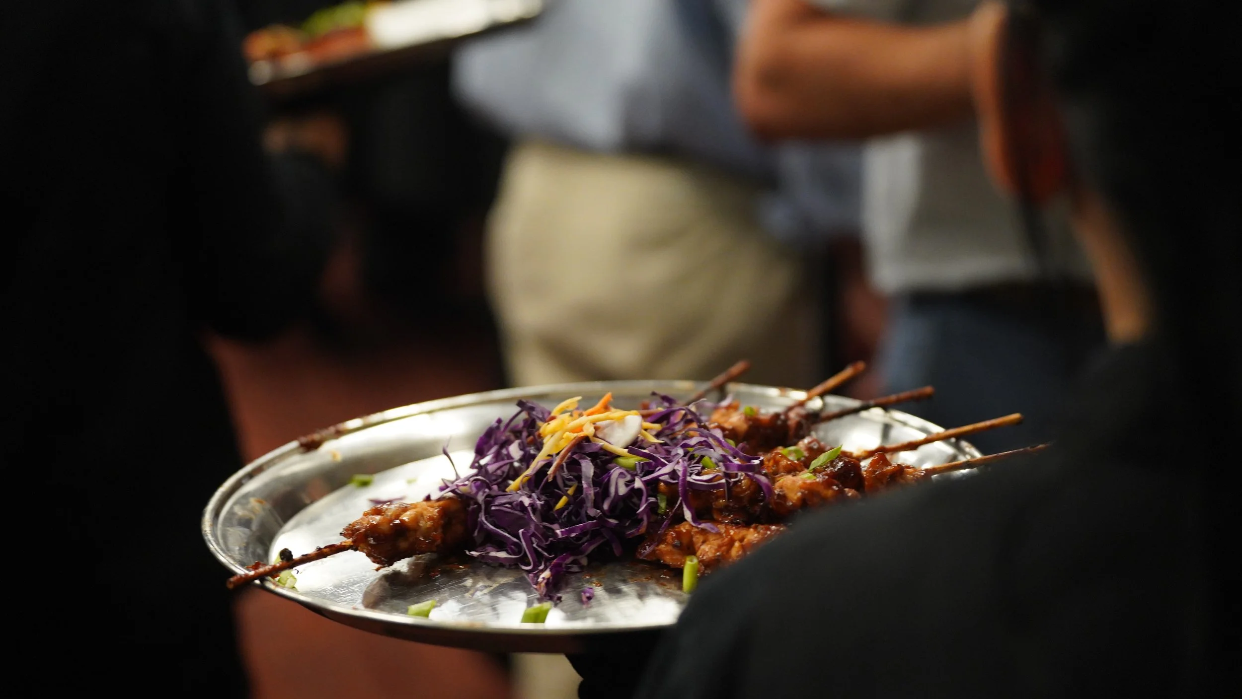 Person serving food with skewers and a colorful salad of purple cabbage and shredded cheese.