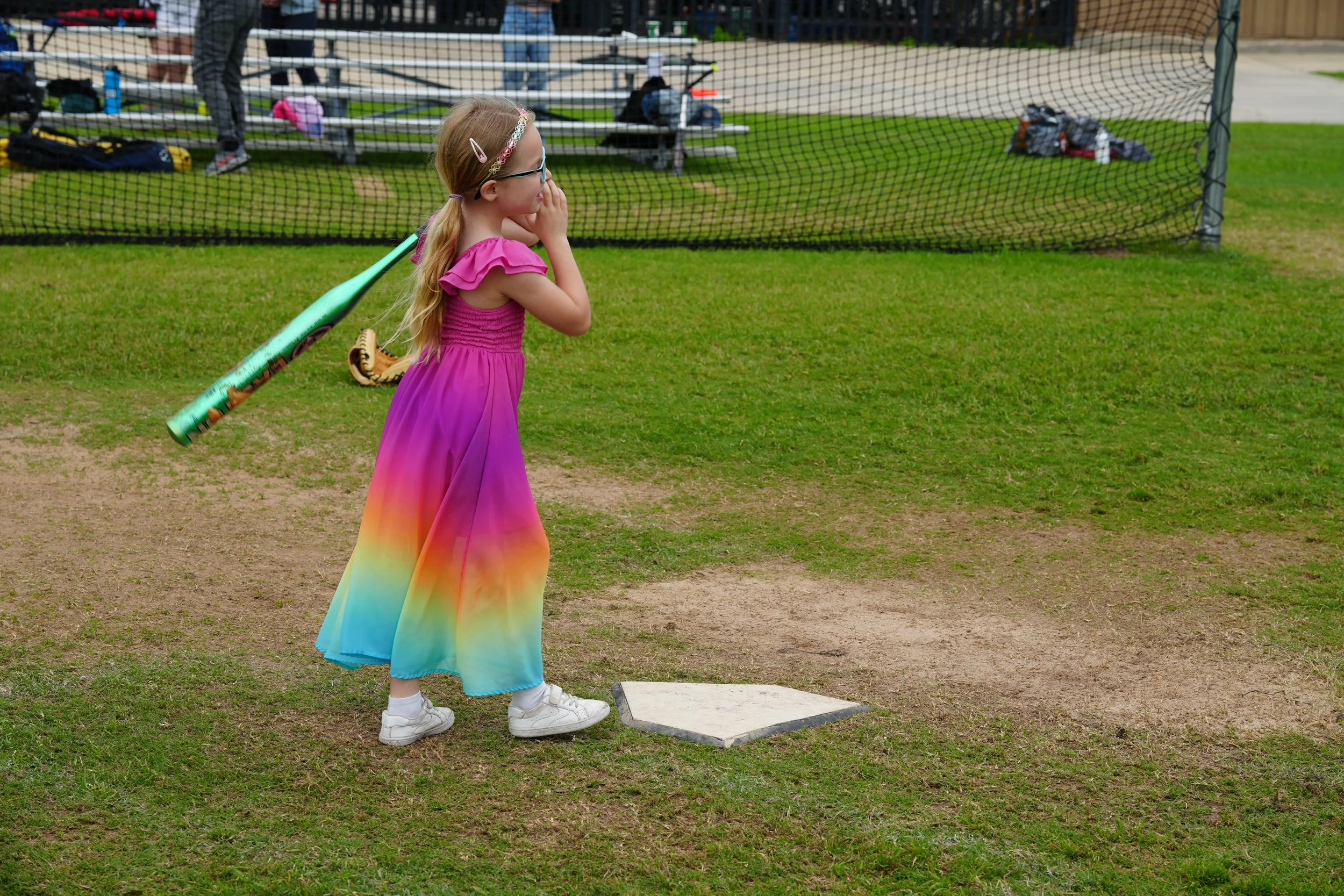 A young girl wearing a colorful rainbow dress, glasses, and a hair accessory, standing on a baseball or softball field near a home plate with her hand covering her mouth, watching a game or practice.