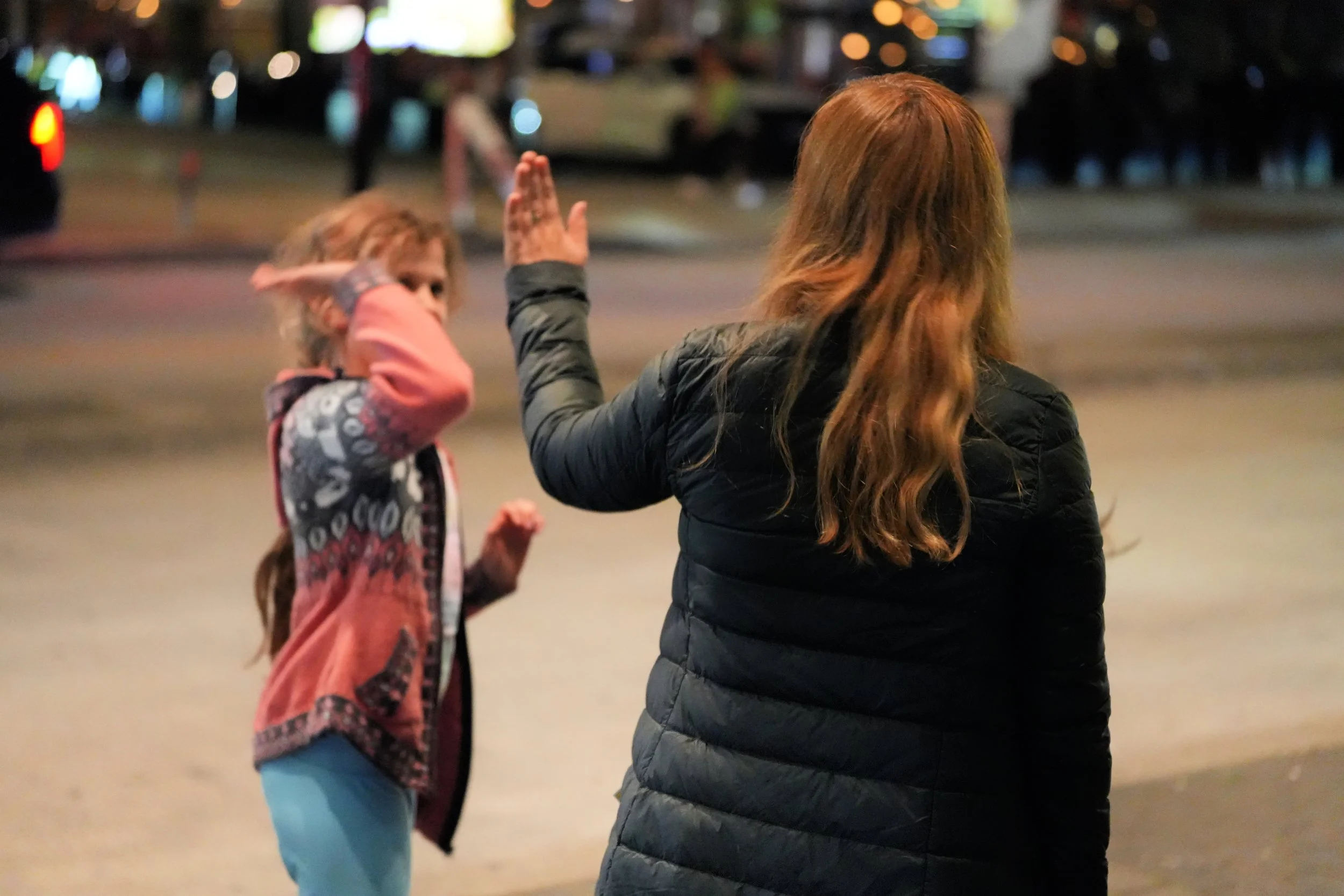 Two females, one with red hair wearing a black jacket, giving a high five in a nighttime outdoor setting, with a young girl in a pink hoodie and blue pants nearby.