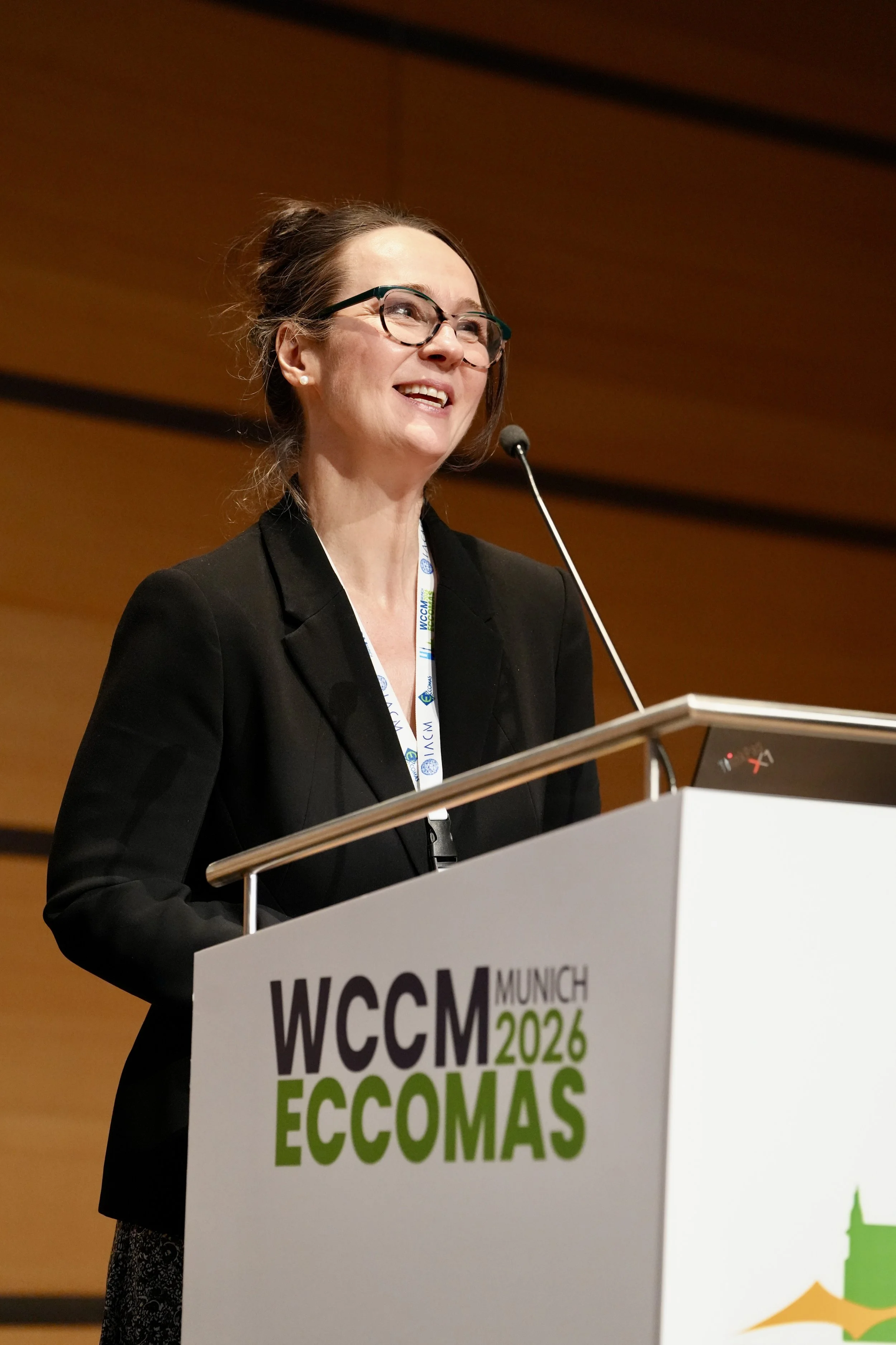A woman with glasses speaking at a podium during an event called WCCM München 2024 ECCOMAS, with a warm background and podium signage.