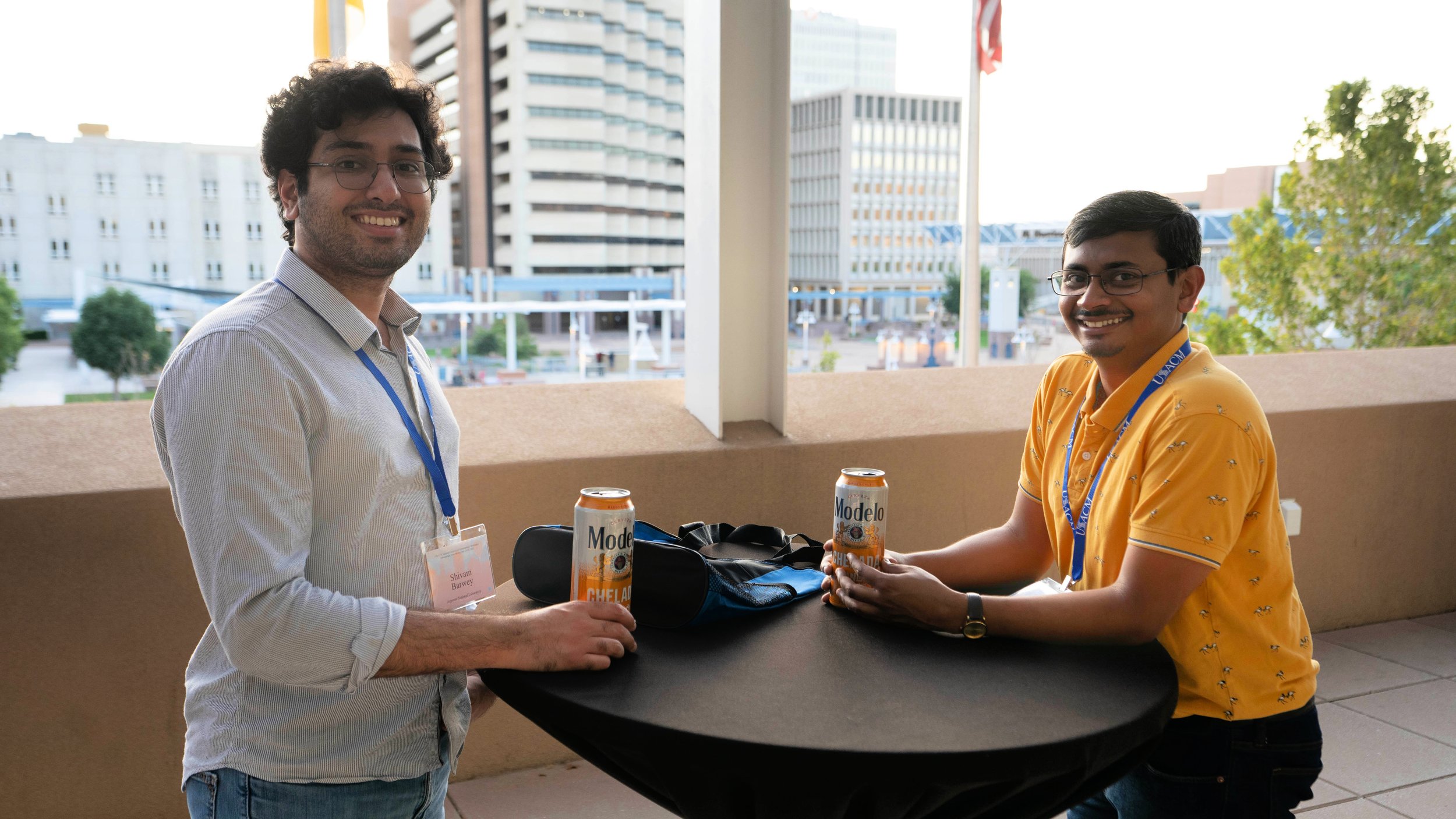 Two men with glasses are smiling and sitting across from each other at a round table outdoors in an urban area. They are holding cans of Modelo Chelada beer. One man is wearing a light-colored shirt with a name tag, and the other is wearing a yellow polo shirt with a pattern and a blue lanyard. There are two other cans of Modelo Chelada beer on the table. In the background, there are tall buildings and some greenery.