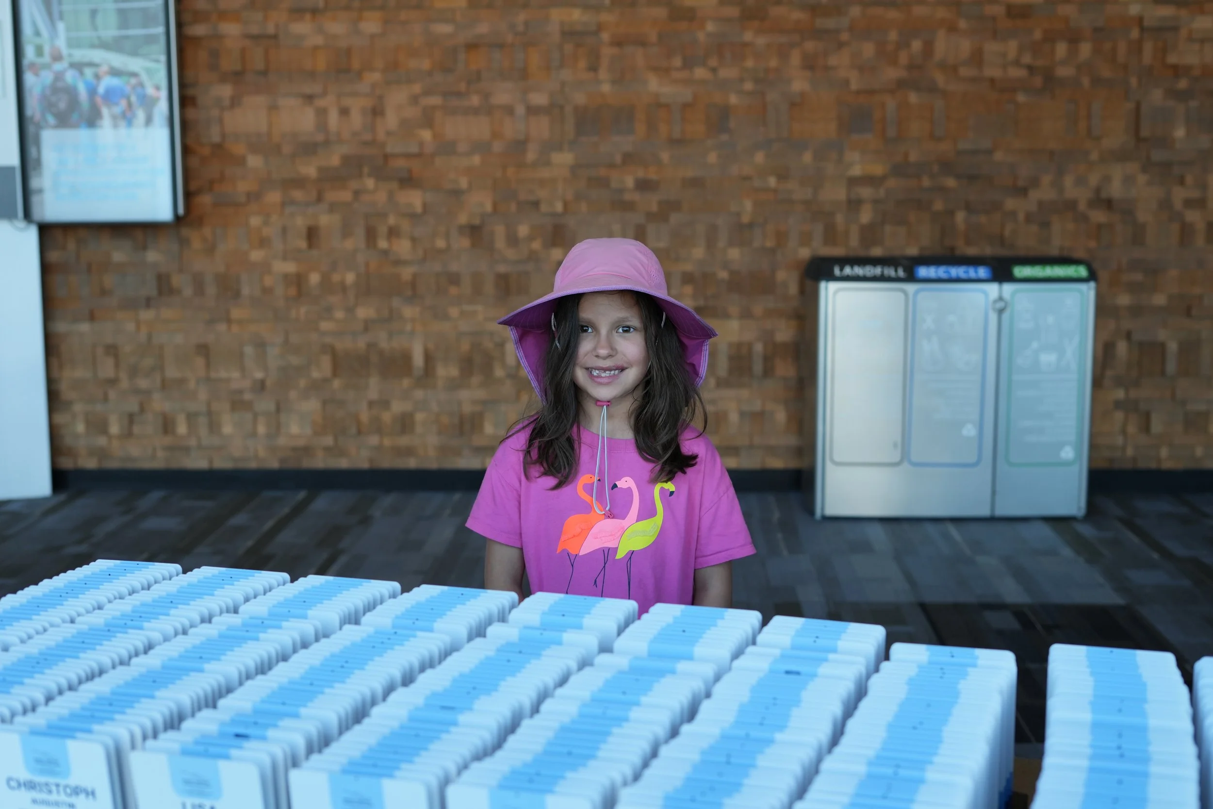 A young girl with long dark hair wearing a pink hat and pink shirt with flamingos stands in front of a display of white and blue medicine bottles.