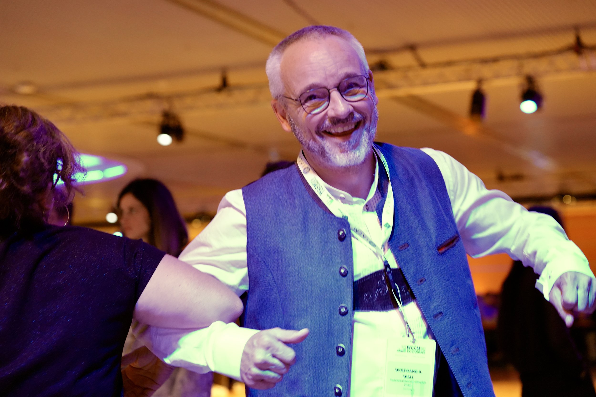 A smiling man with glasses and a gray beard wearing a traditional vest and a conference badge over a lit, at a conference dinner.