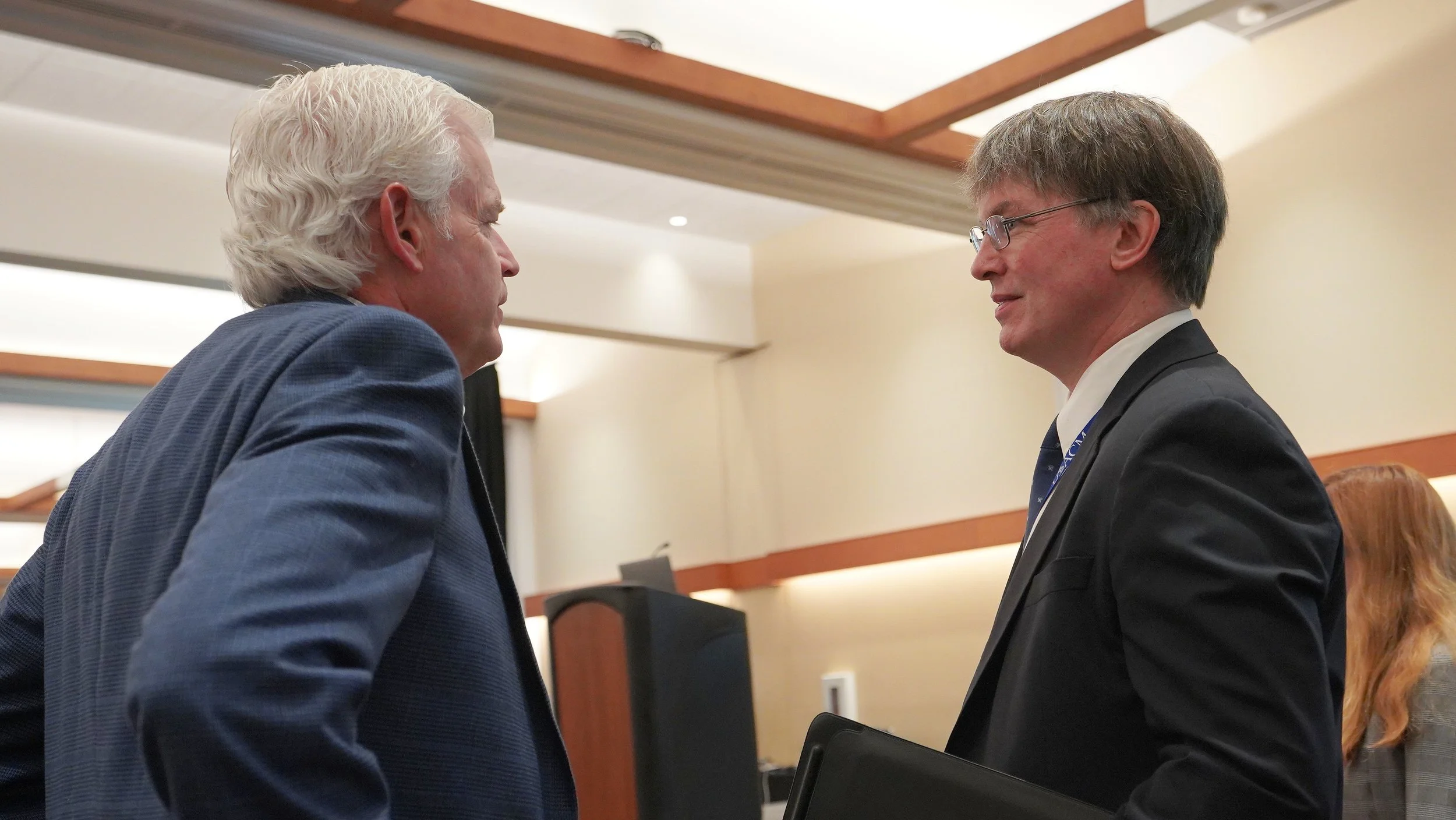 Two men with gray hair wearing suits and glasses are face to face in a professional setting.