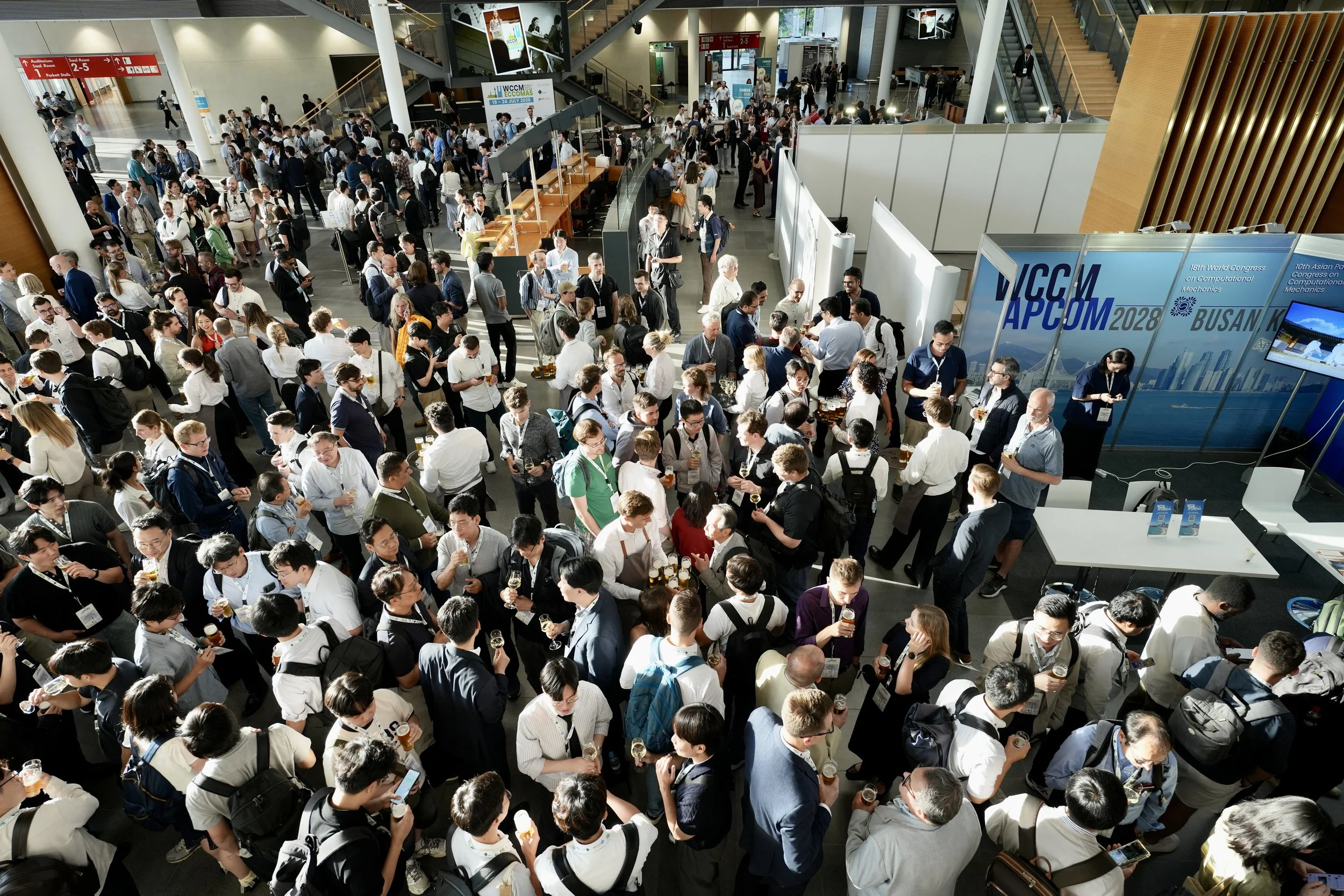 Crowd of people at a conference or event in a large, modern indoor venue with stairs, booths, and digital screens.