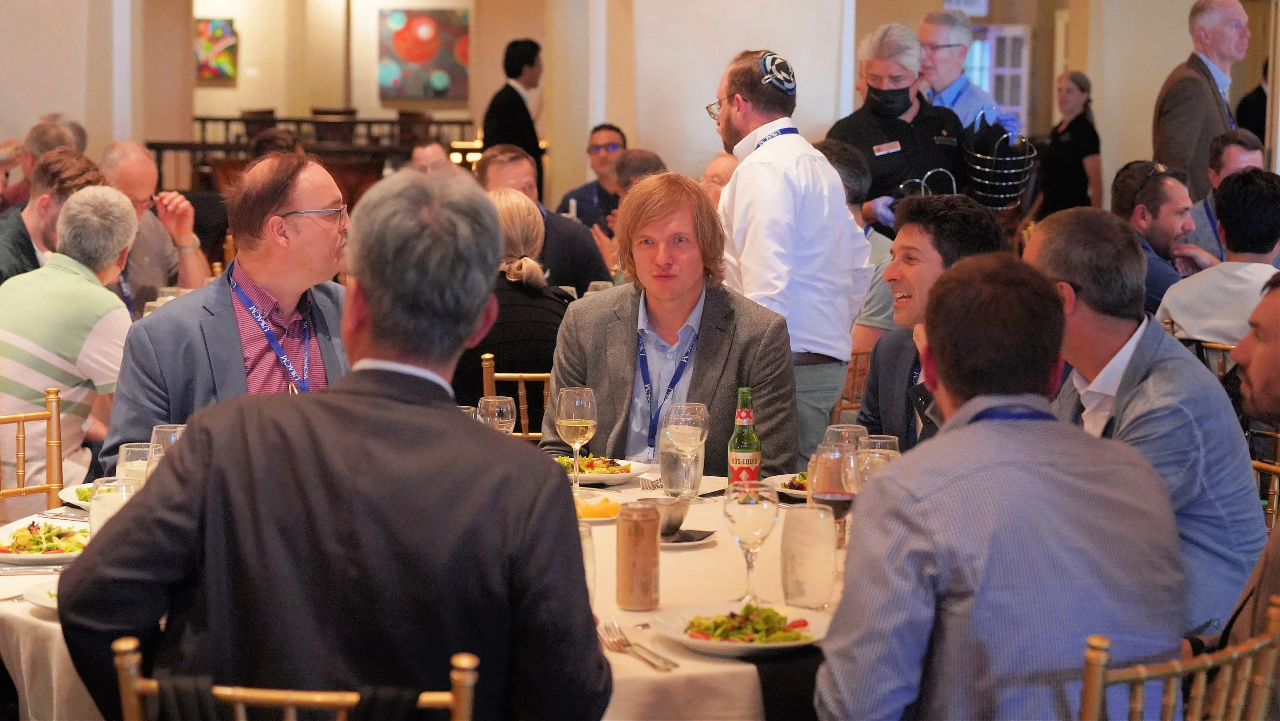 A group of people seated at a formal dinner, engaged in conversation, with several glasses and plates of food on the table, and a waiter serving in the background.