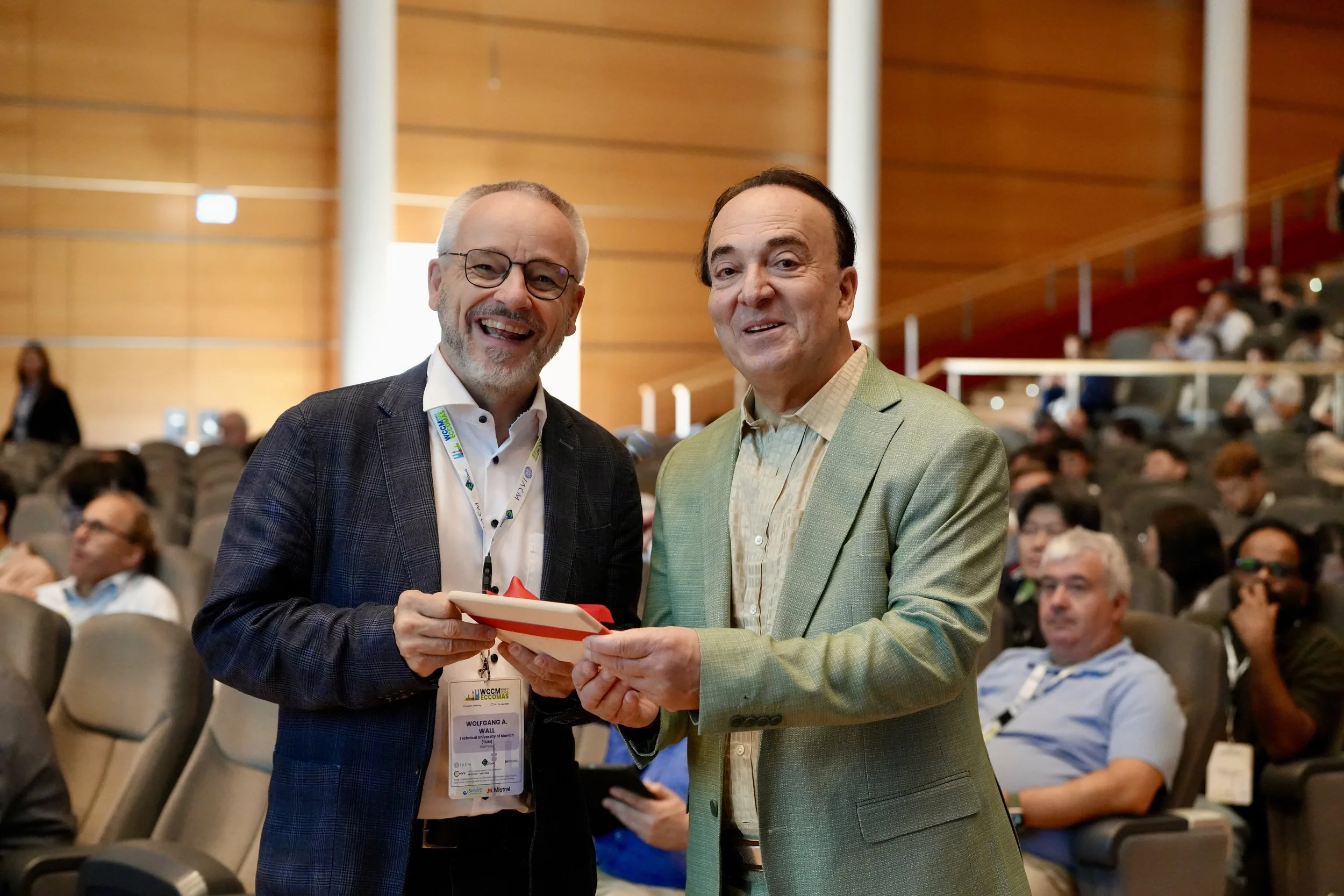 Two men in business attire exchanging an award or certificate at an academic conference or event, with many people seated in the background.