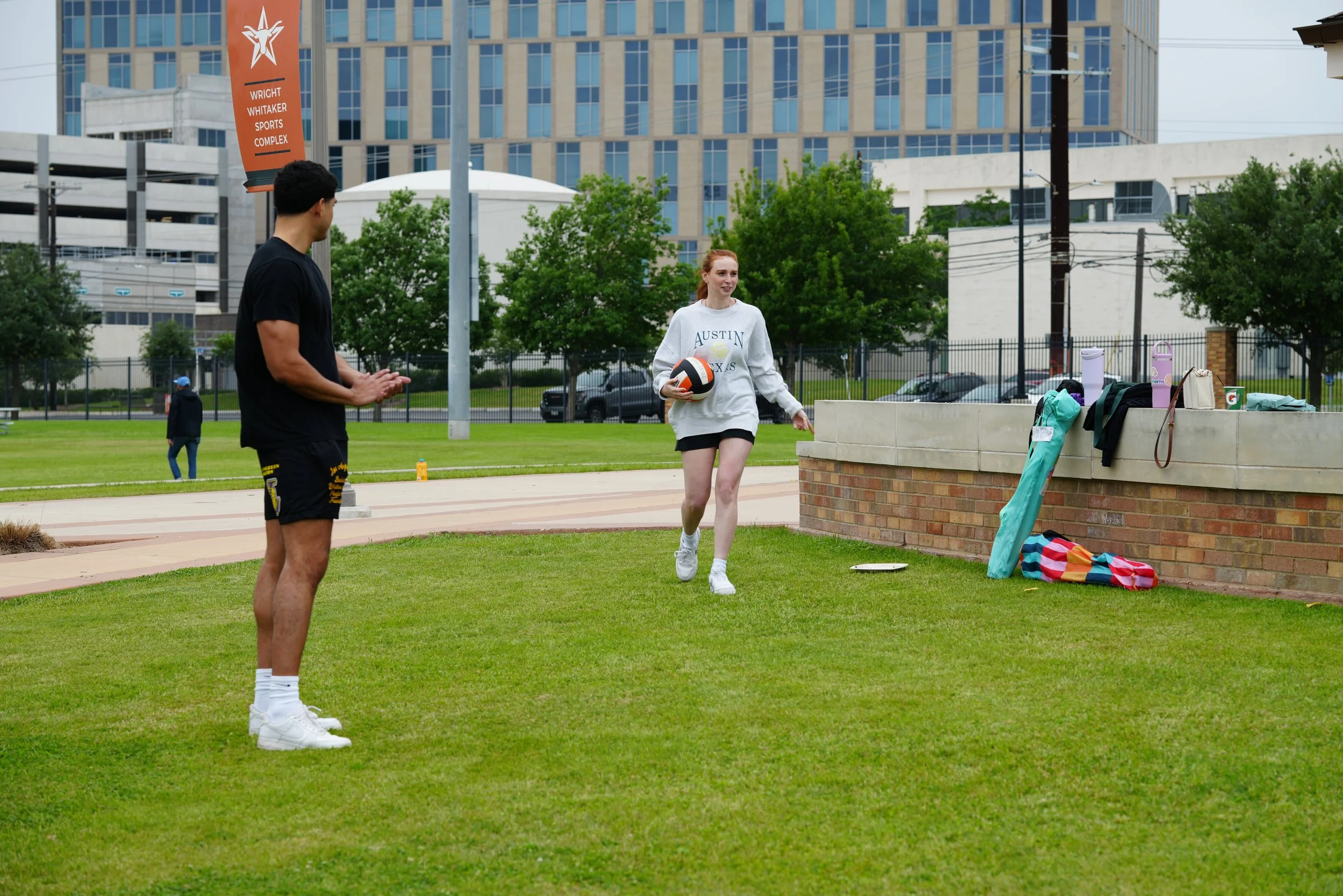 A young woman with red hair holding a volleyball and smiling, standing on a grassy field next to a brick wall with various personal items on top, while another young man in black sportswear stands nearby. In the background, trees, a parking lot with cars, and a tall building with blue glass windows are visible, suggesting an outdoor sports or recreation area in an urban setting.