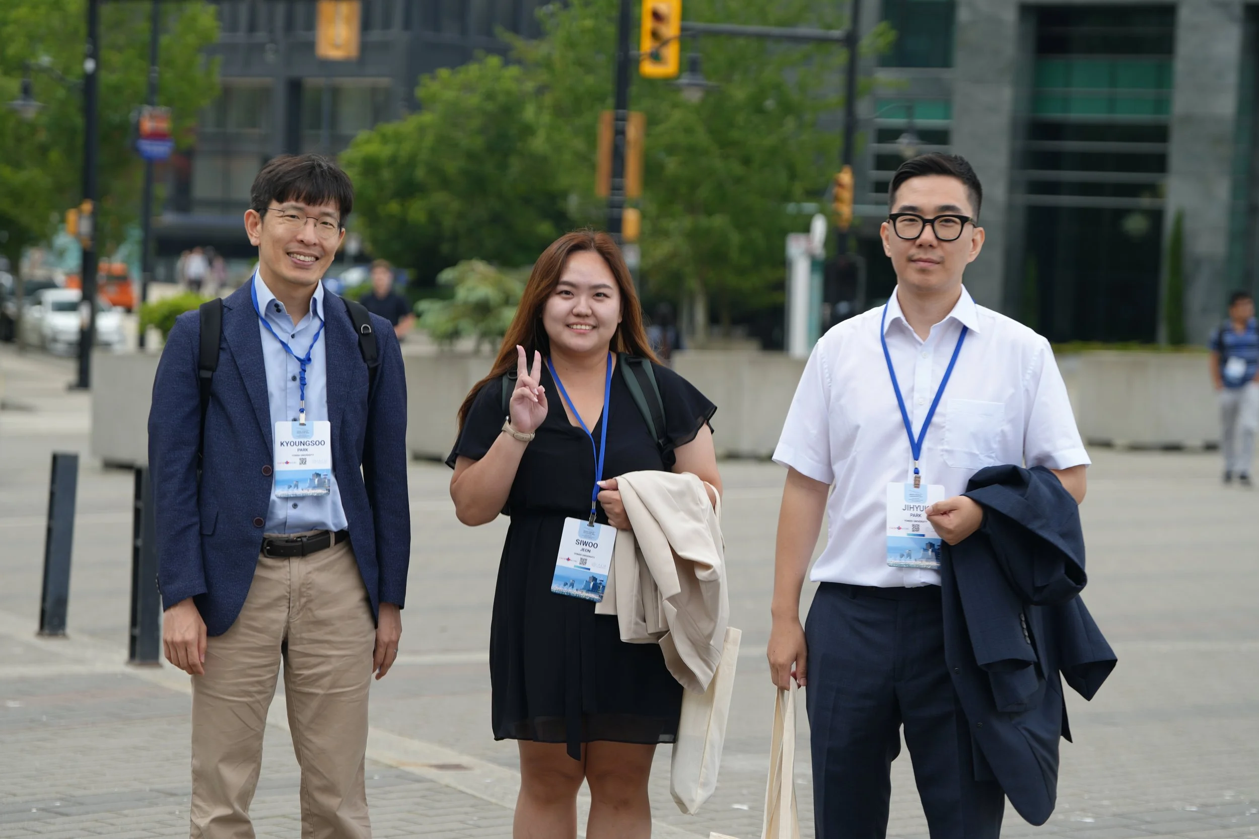 Three young adults, two men and one woman, standing outdoors in an urban setting with modern buildings and trees in the background. They are smiling, and the woman is making a peace sign. All are wearing conference badges on blue lanyards.