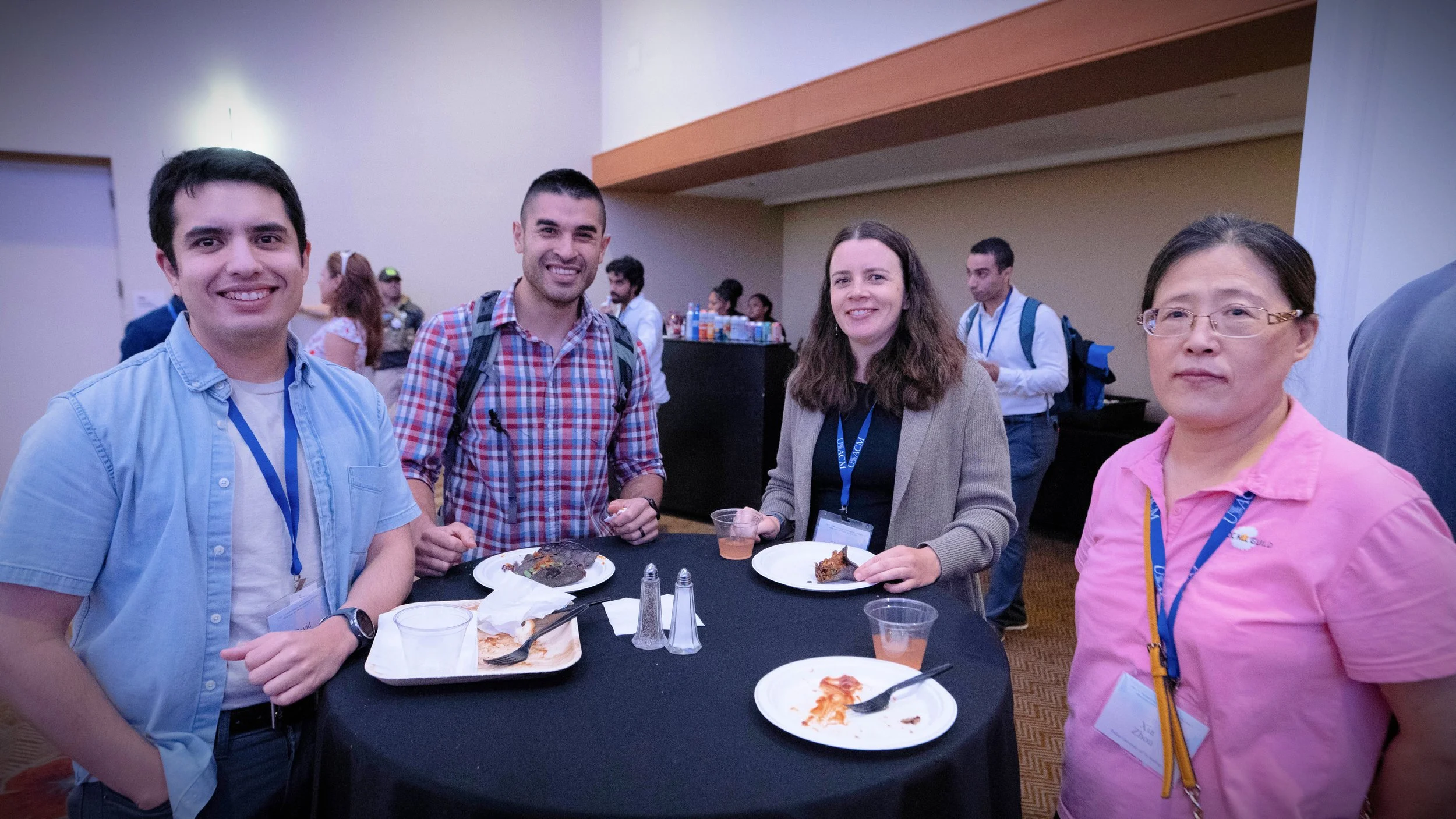 Group of four people standing around a table with food and drinks at a conference or event, smiling for the camera.