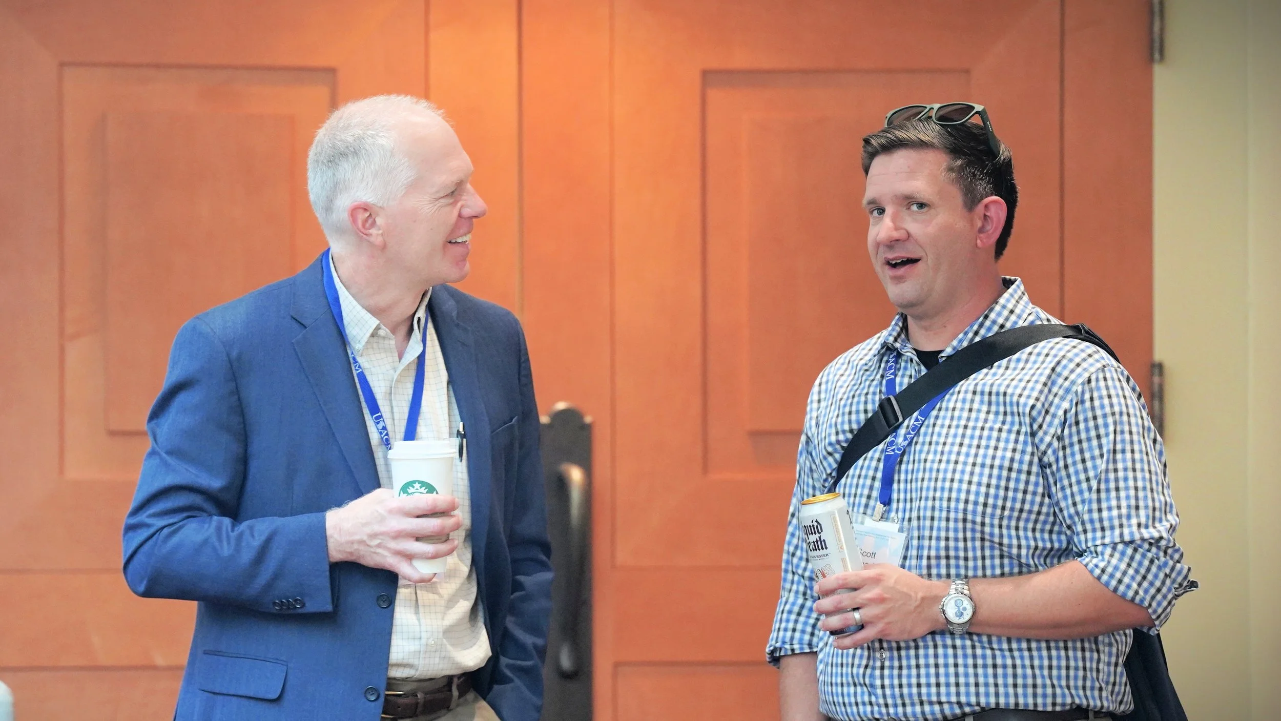 Two men are standing and talking at an event. The man on the left is wearing a blue blazer and holding a Starbucks coffee cup, while the man on the right is wearing a checked shirt, sunglasses on his head, and holding a can of Liquid Death water. They are both wearing conference name tags and are in front of a wooden-paneled wall.