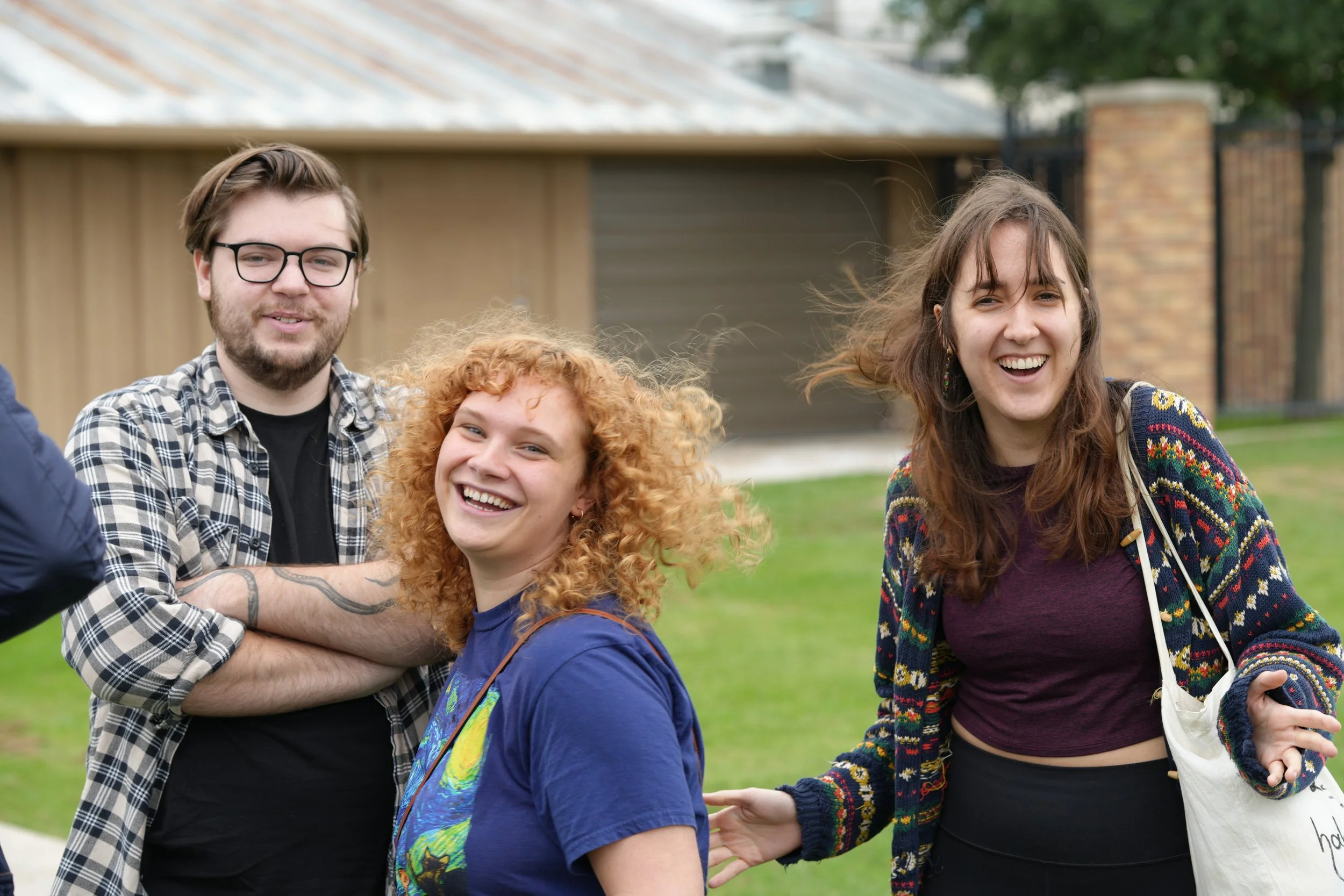 Group of four diverse young people smiling and standing together outdoors near a brick and wood fence, with green grass and trees in the background.