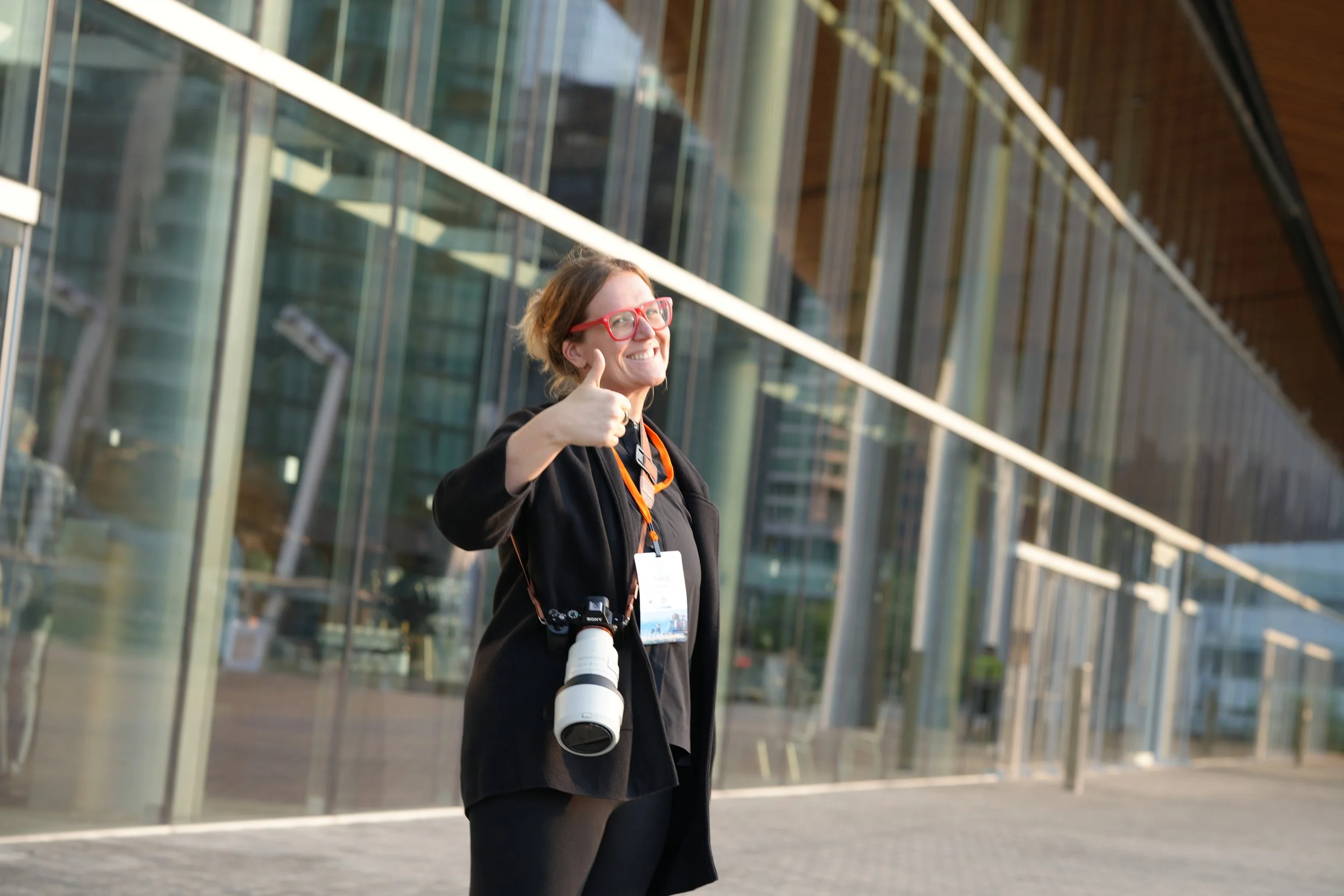 A woman with red glasses giving a thumbs-up outside a modern glass building, wearing a black jacket with a camera hanging around her neck.
