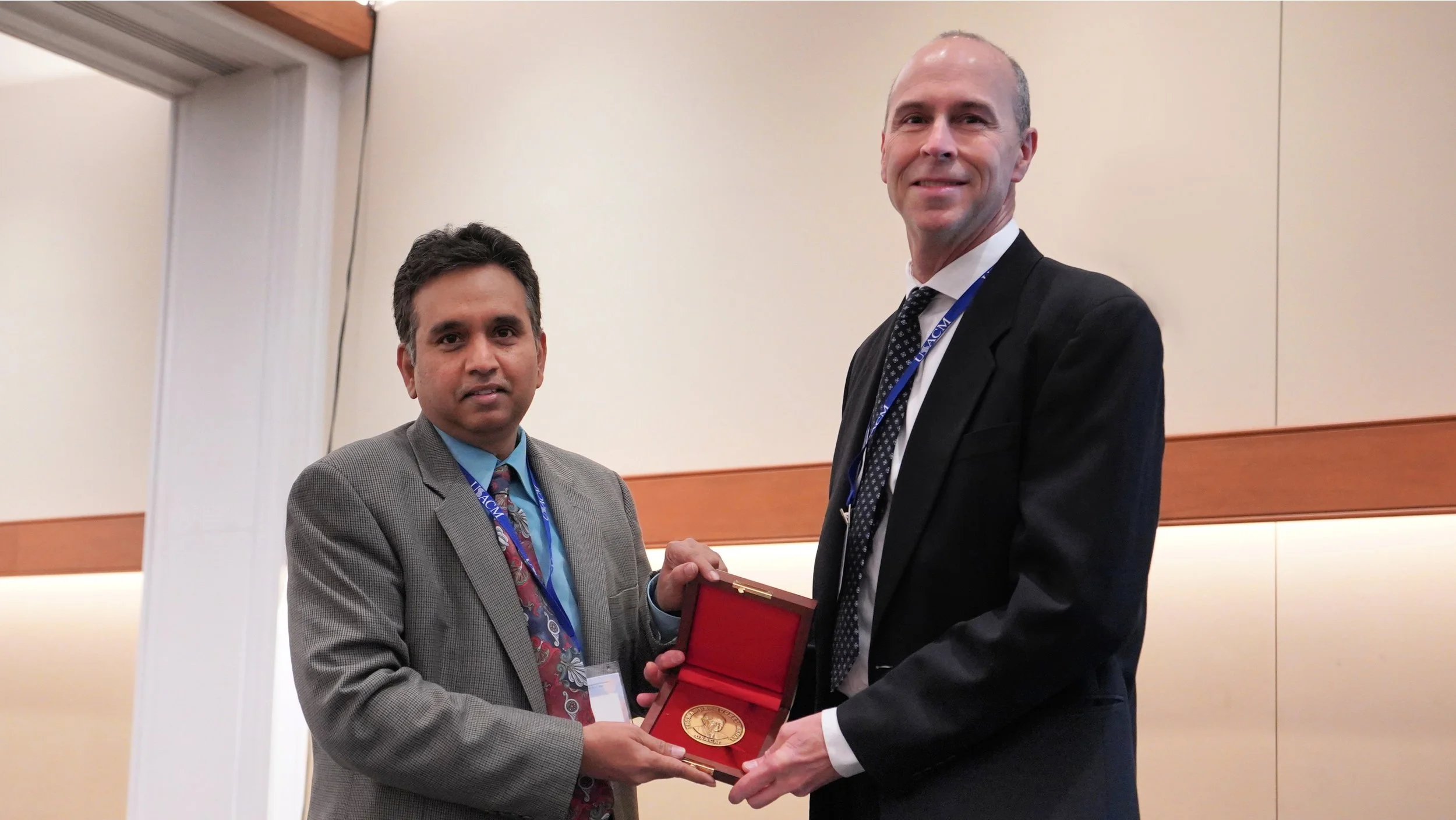 Two men in suits shaking hands during an award ceremony, one man is handing a medal to the other in a conference room.