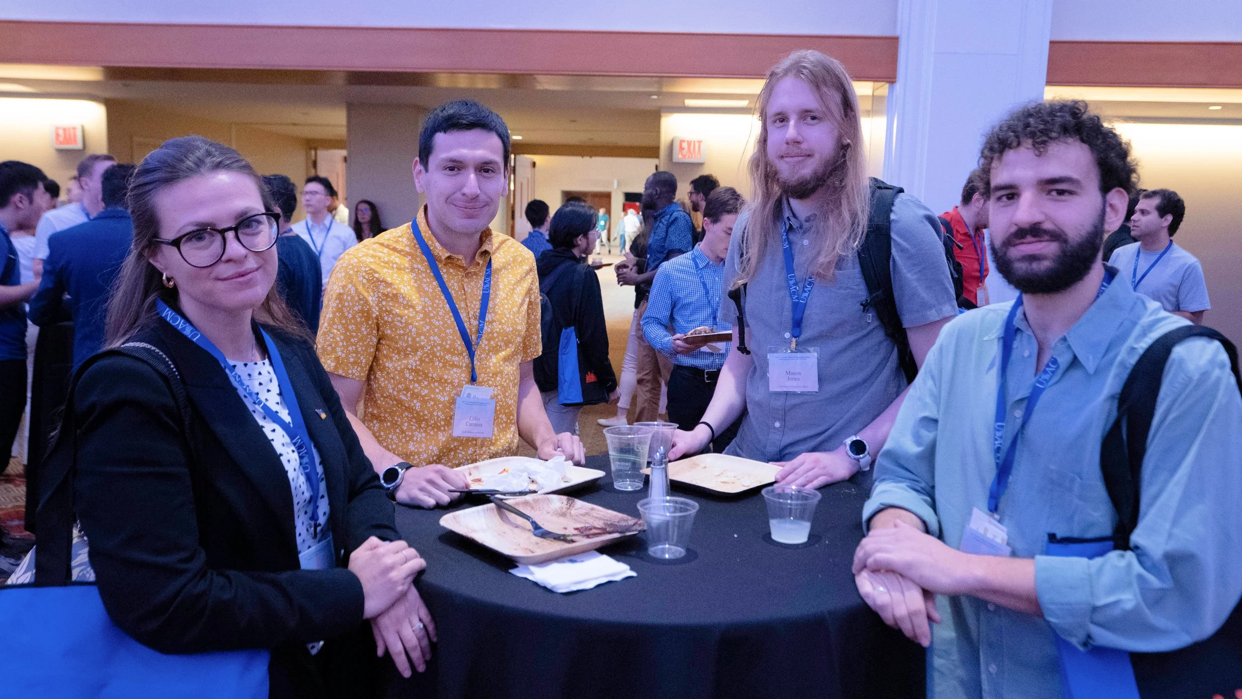 Group of four young adults at a conference or networking event, sitting at a table with food and drinks, wearing conference badges and lanyards, with many other people in the background inside a large hotel or conference center.