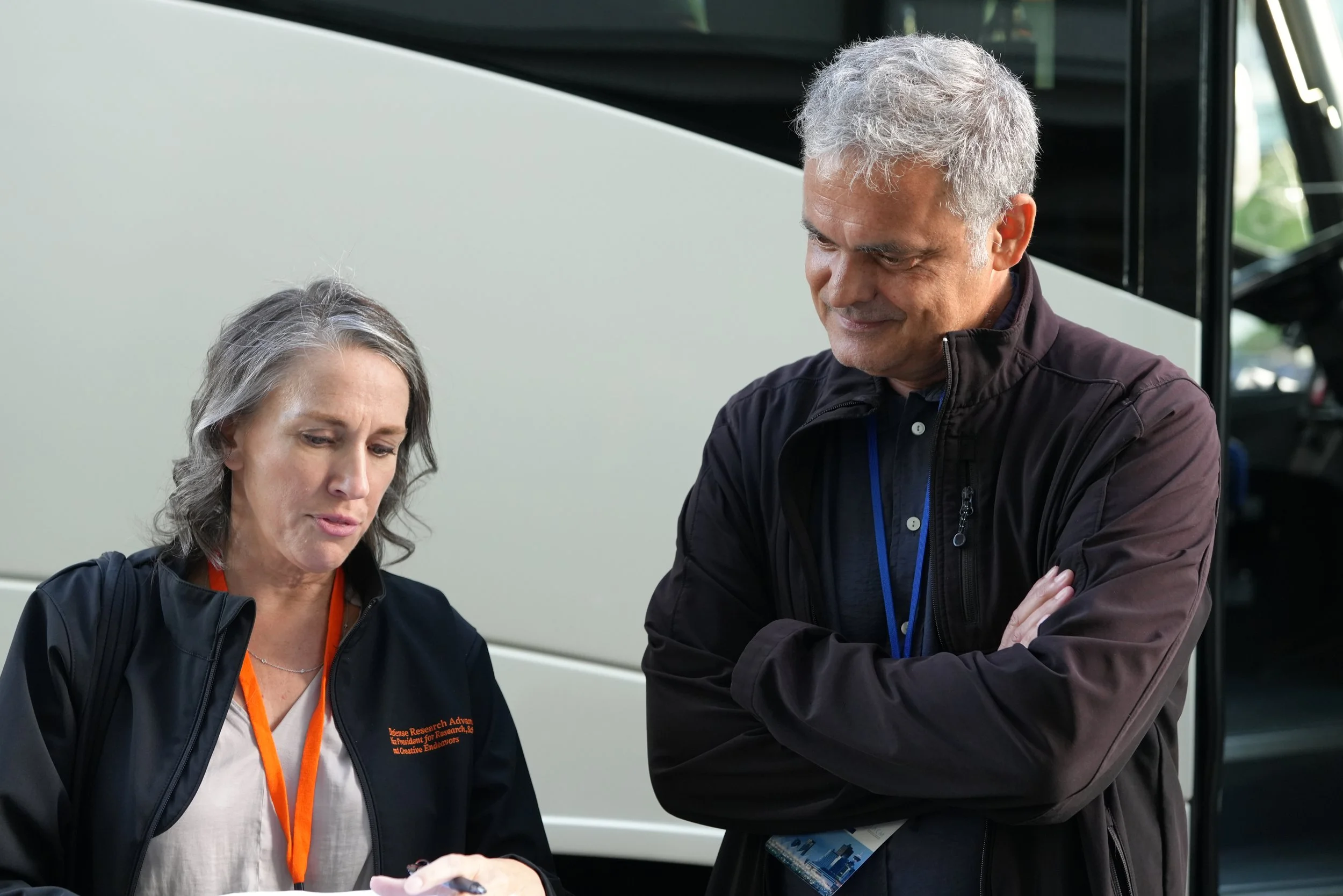 Two middle-aged adults, a woman and a man, are engaged in a conversation outdoors near a vehicle. The woman has shoulder-length gray hair, and the man has short gray hair. Both are wearing black jackets, and the woman has a white shirt underneath. They seem to be examining something together, with the woman holding a pen and the man with his arms crossed. The background shows part of a bus or large vehicle and some greenery.