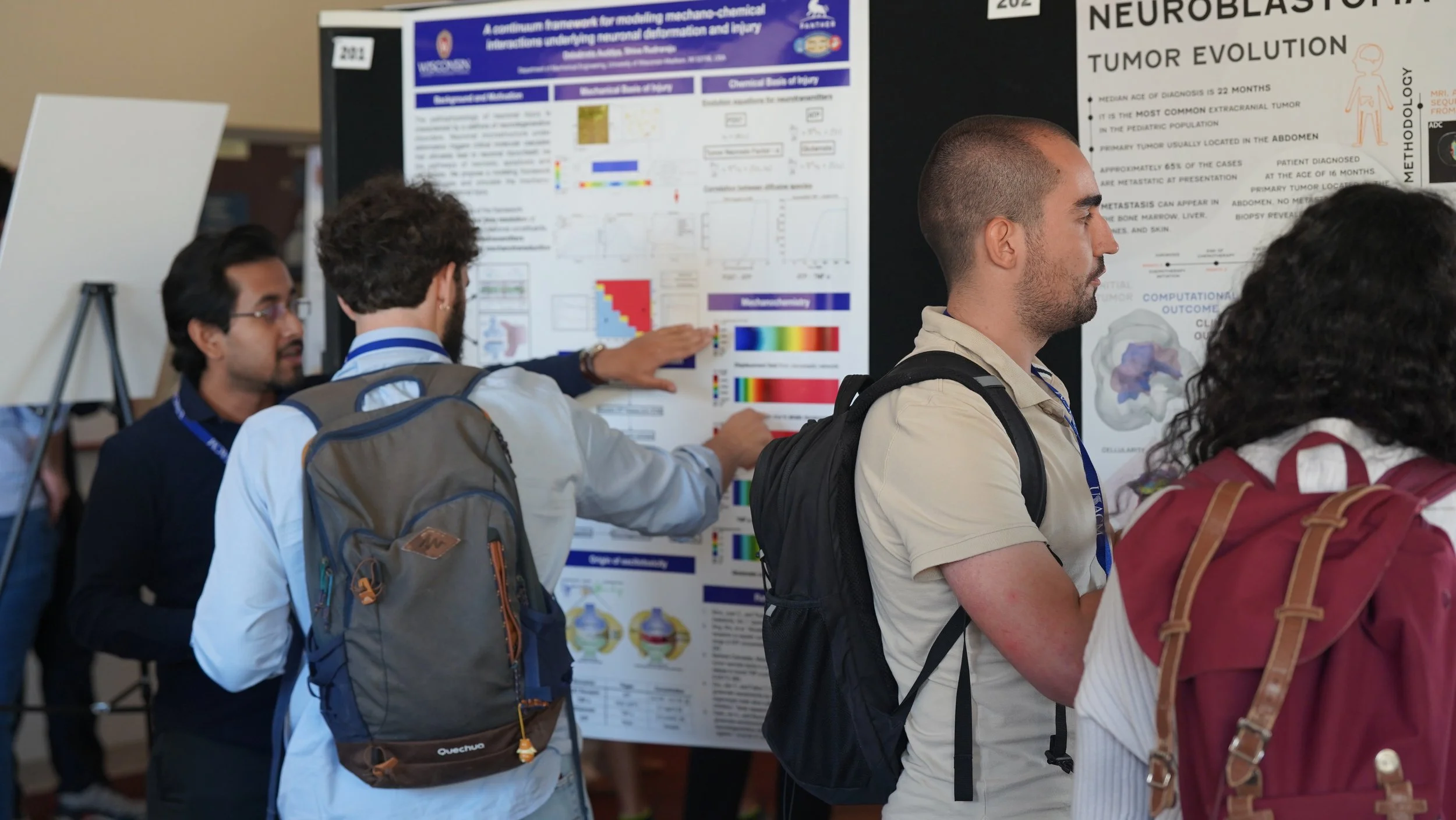 A group of people, some with backpacks, are viewing scientific research posters at conference. One man in beige shirt and others are engaged in discussion around the posters.